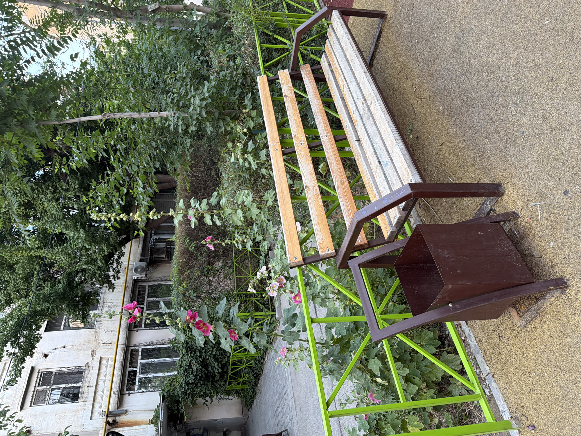This is an outdoor shot of a park bench and surrounding greenery. The bench has a dark brown metal frame and wooden slats for the seat and backrest. Attached to the side of the bench is a dark brown, cubical trash receptacle. The bench is situated next to a patch of green foliage and some blooming pink flowers. Behind the foliage, a building with multiple windows is visible. The building appears to be aged, with some visible wear on its exterior. Above the building, trees with dense green leaves can be seen. The ground around the bench is paved with a textured, yellowish material. The image is oriented vertically, suggesting it was taken with a phone camera. The lighting suggests it is daytime, likely with overcast conditions due to the diffused light. The scene appears to be a quiet public space, possibly a courtyard or a small park area. There are no people or animals visible in the image. There is also no visible text.