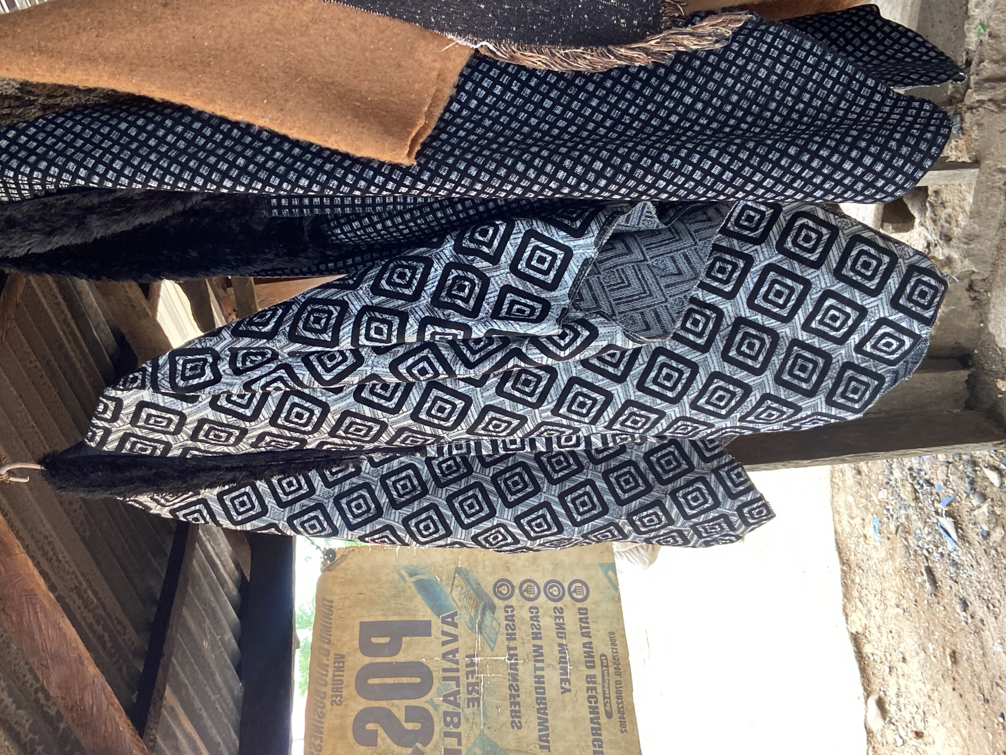 In Mararaba, Nigeria, at a roadside stall under a corrugated metal roof supported by rough wooden beams, several garments hang from hooks. The most prominent is a light-colored jacket with a black geometric diamond pattern. To its right, a black garment with a white checkered pattern is partially visible, and further right, a tan garment with frayed edges hangs. In the background, a weathered sign displays text and an image of a POS machine, indicating business services such as cash transfers and sending money. The ground beneath the stall is a mixture of sand and small stones.