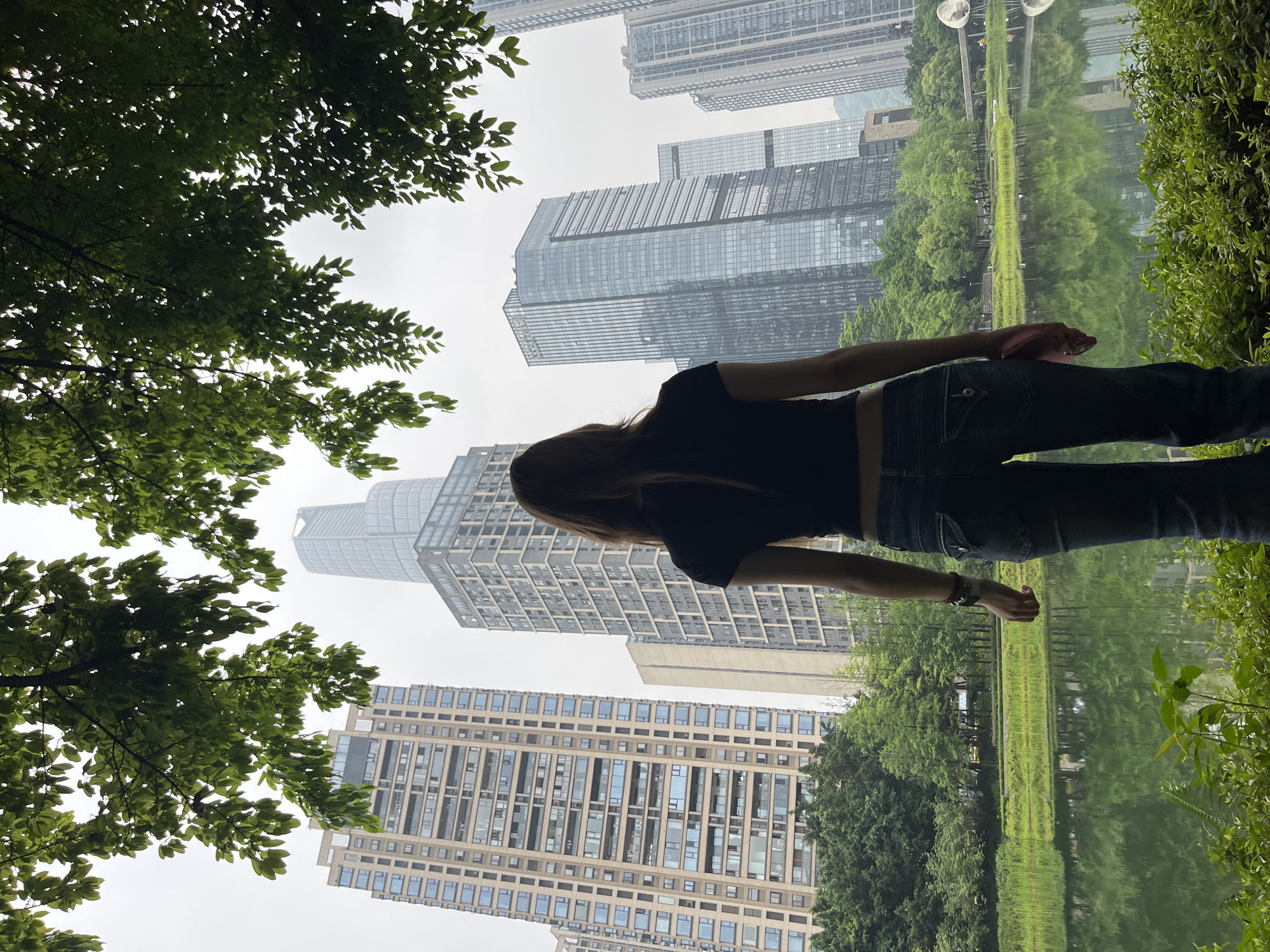 A woman stands with her arms outstretched near a body of water in Yuzhong District, China. Her dark hair flows down her back, and she is wearing a black top and dark jeans. Tall buildings, including skyscrapers, form a backdrop, some partially obscured by trees. The water reflects the surrounding buildings and trees. Tree branches with green leaves frame the top left corner of the image. The sky is overcast and hazy.