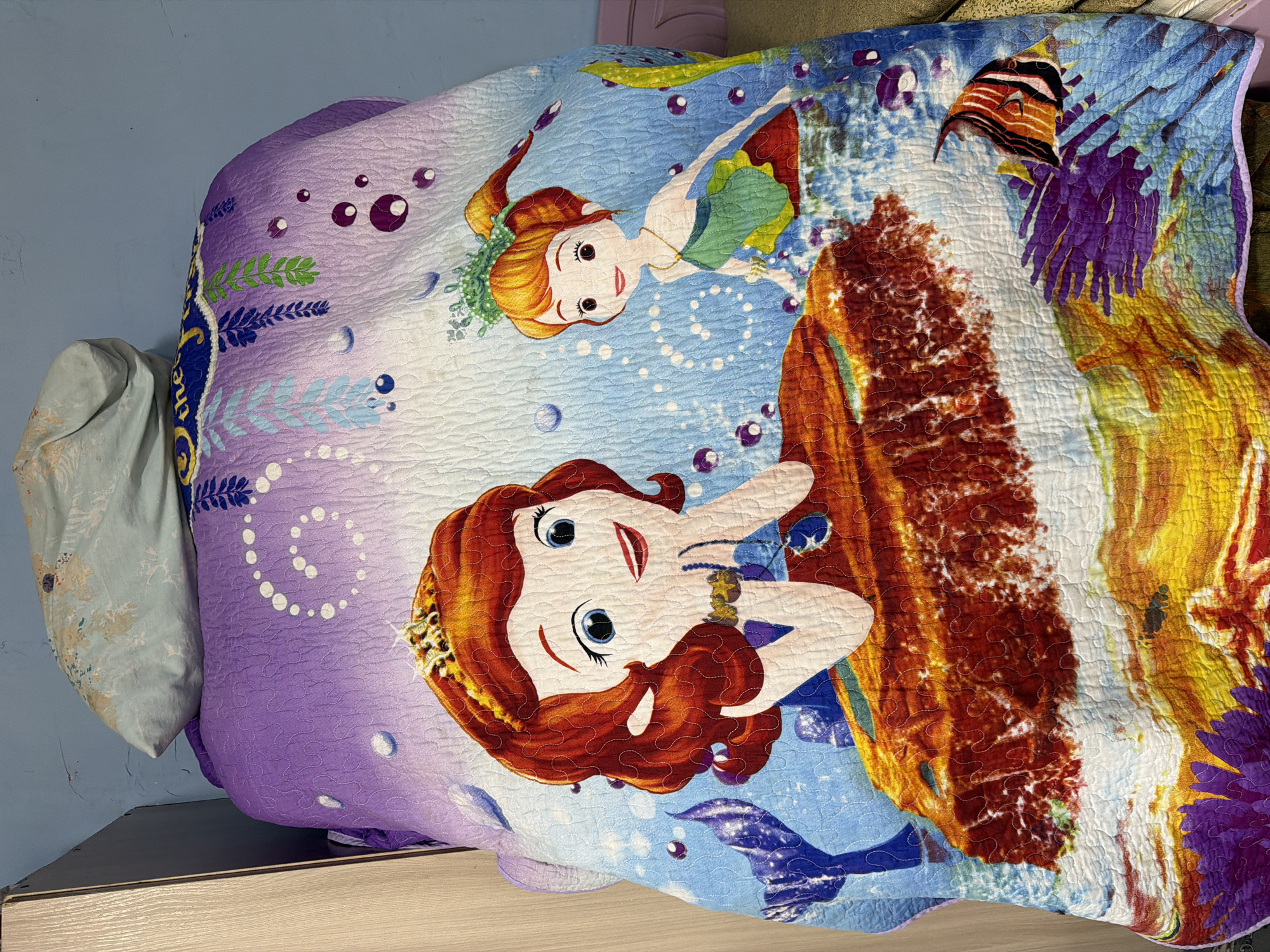 A close-up, eye-level shot shows a colorful quilt draped over a bed in Köhnə Quşçu, Azerbaijan. The quilt features an underwater scene with two cartoon mermaid characters. The larger mermaid, on the left, has long red hair and a golden crown, and is leaning on a rock formation with a jewel-toned necklace. The smaller mermaid, on the right, has orange hair pulled back in a ponytail, wears a green top, and has a leafy crown. The background of the quilt is a vibrant blue with bubbles and purple and green seaweed. A pillow with a blue and white floral pattern is placed on top of the quilt, partially obscuring the top of the bed. The room appears to be dimly lit, and the wall behind the bed is painted light blue. To the left of the bed is a sliver of a wooden wardrobe. The bottom of the quilt reveals a detailed depiction of coral and a tropical fish.