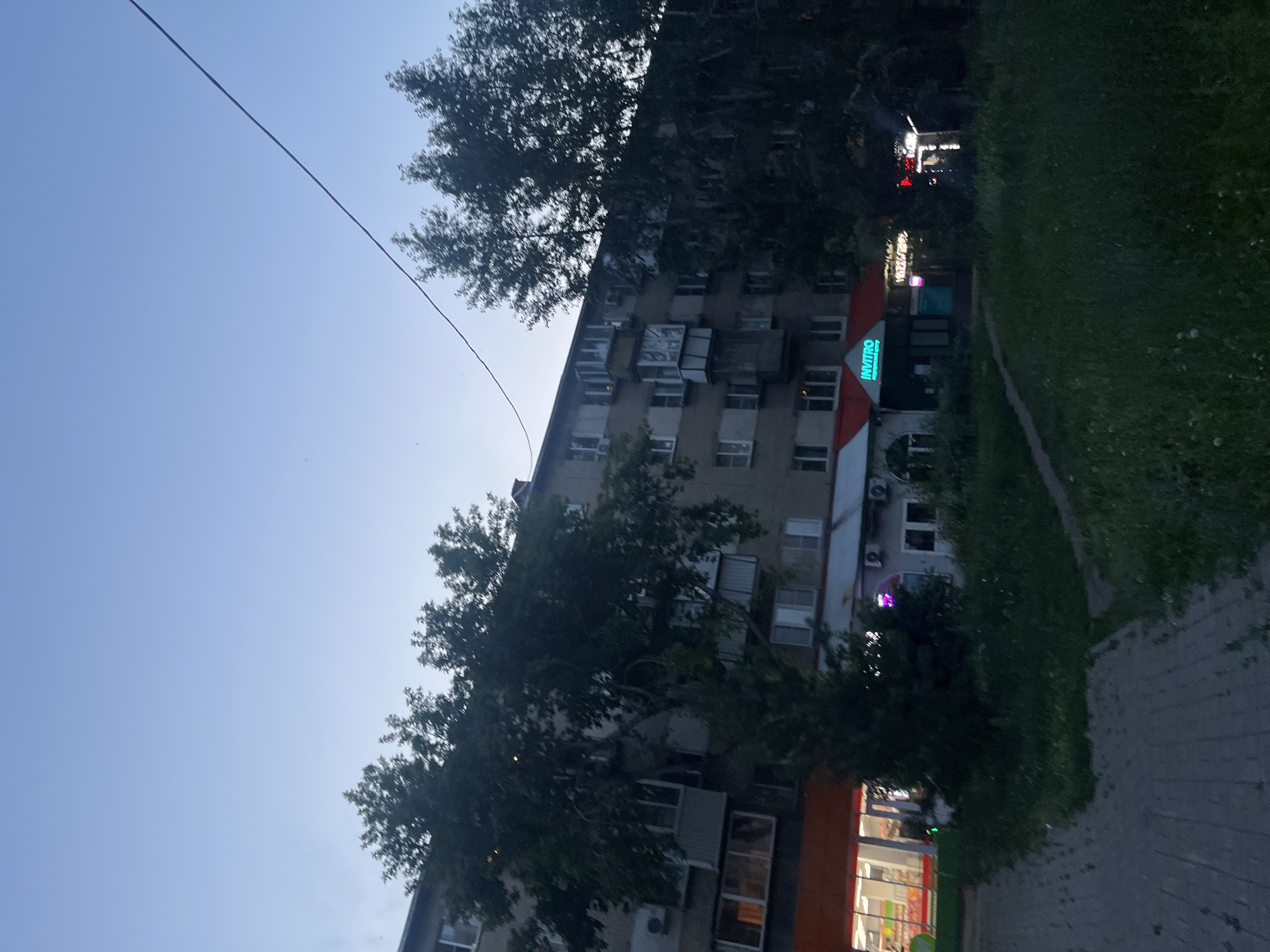The image depicts a street-level view of a residential building in Chelyabinsk, Russia, likely during twilight or early evening. The building is multi-story with several windows and balconies visible. Trees with dense foliage partially obscure the building, casting shadows on its facade. In front of the building, there are businesses with illuminated signs, including one that reads "INVITRO" in bright green neon letters, suggesting a medical laboratory or clinic. Another sign with red lettering is also visible. The ground in front of the businesses is paved, with a path leading through a grassy area. The sky above is a gradient of light blue, indicating the transition from day to night.
