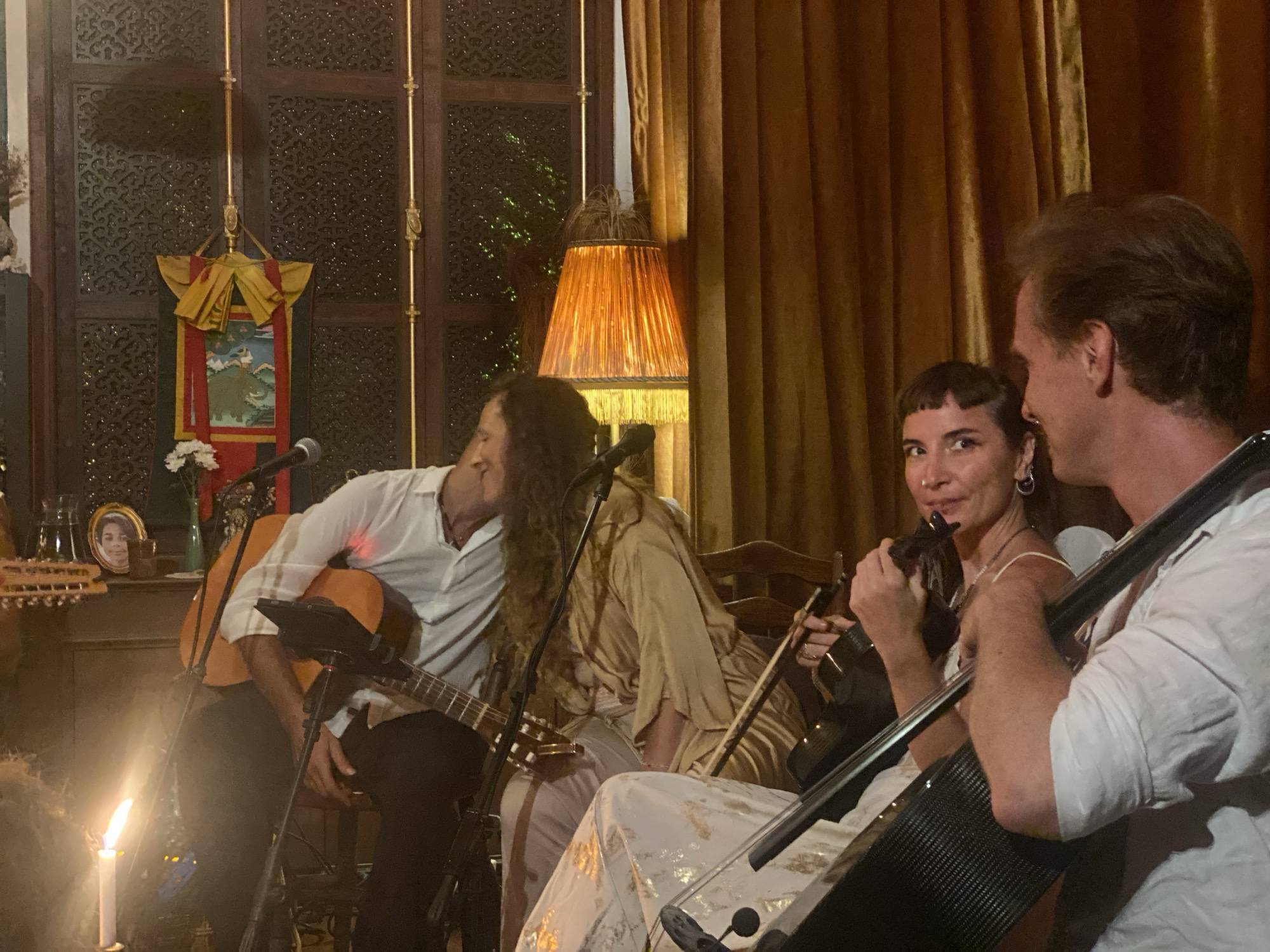The image captures a live music performance, likely in an intimate setting, possibly a restaurant or bar, in ตำบลเกาะพะงัน, Thailand. Three musicians are visible: one playing an acoustic guitar, another a violin, and the third a cello. The guitarist, wearing a white shirt and dark trousers, is seated. The violinist, also in white, has a short haircut and is looking towards the camera. The cellist, wearing a white shirt, is positioned on the right side of the frame, partially facing away. An additional person, dressed in tan, is seated between the guitarist and violinist. The room is lit with warm, dim light from lamps, and a candle provides additional illumination on the left. There is a decorative wooden screen and a framed photo on the left side. The background features golden curtains.