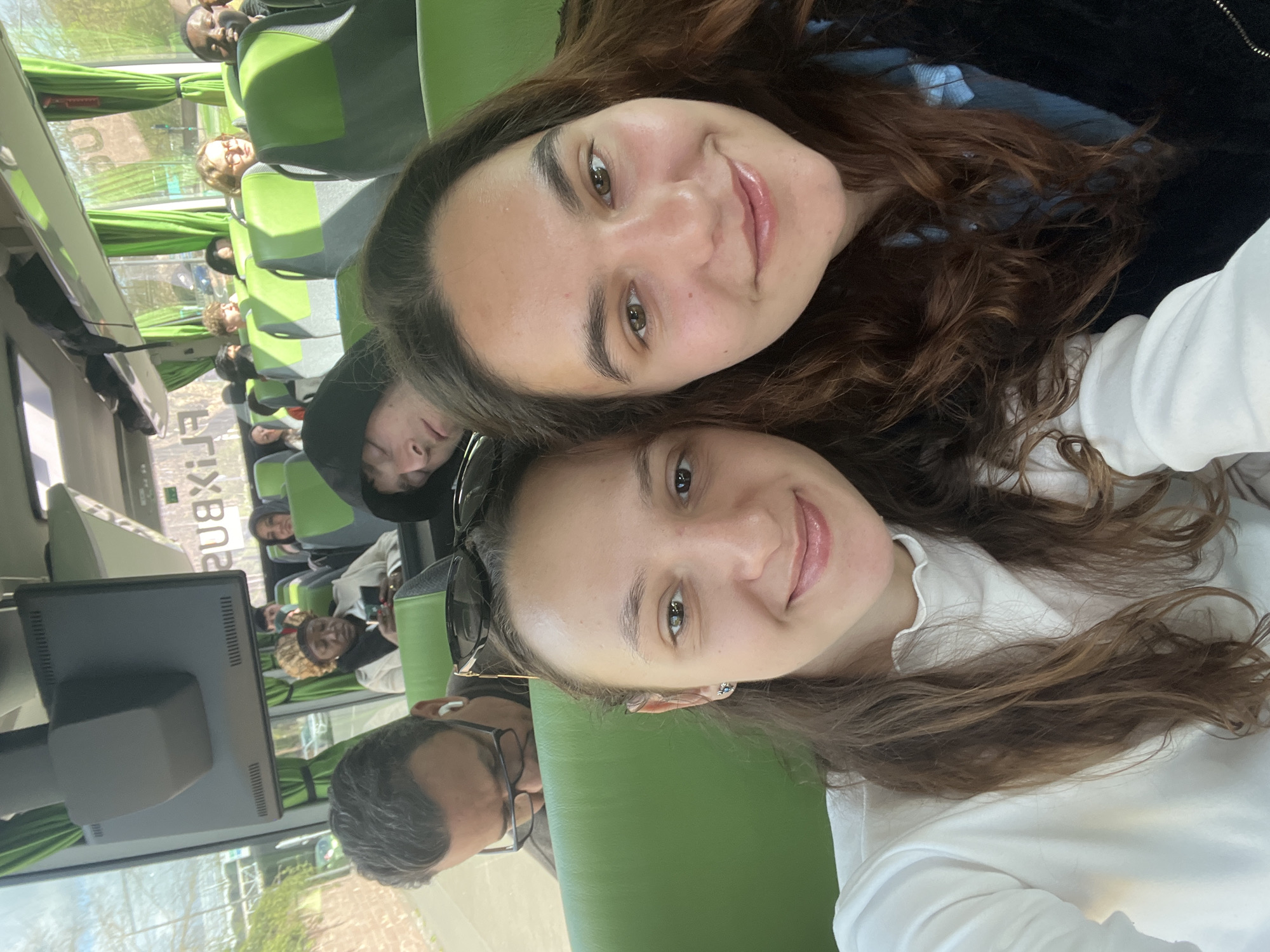 Two young women are posing for a selfie. They are seated in a vehicle, likely a bus, with other passengers visible in the background. The interior of the vehicle has green seating. Outside the windows, building structures with writing in the windows are visible. This is potentially the city of Metz, France. Other passengers are visible in the background of the bus. A man with glasses is to the bottom left of the frame.