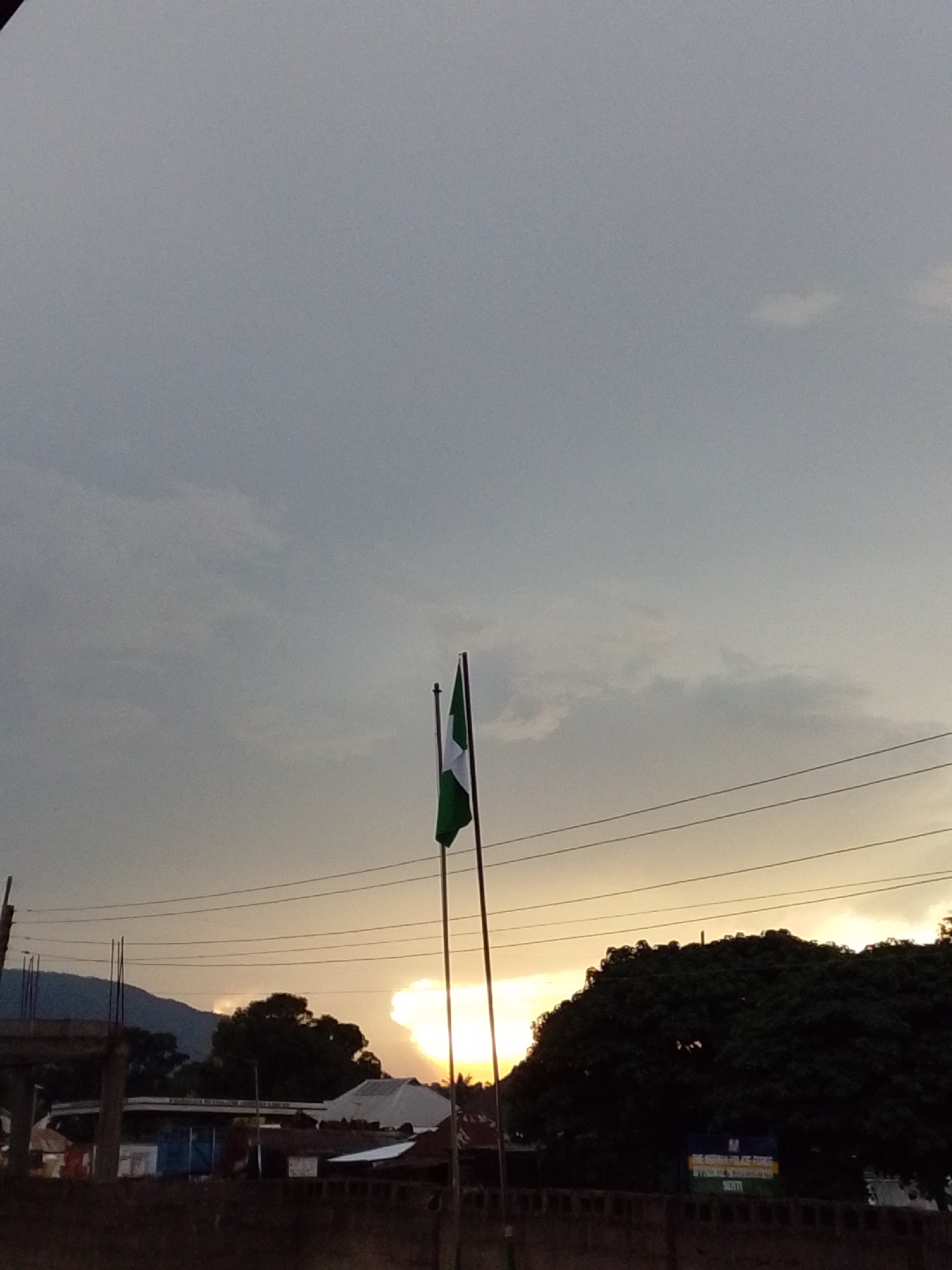The image depicts a twilight scene in Serti, Nigeria, with a Nigerian flag prominently displayed on a flagpole. The flag, a vertical triband of green, white, and green, is slightly furled, suggesting a gentle breeze. The sky is overcast with a soft grey hue dominating, but the setting sun is breaking through the clouds, casting a warm, golden glow towards the horizon. This creates a dramatic contrast between the bright sky near the sun and the darker, muted tones of the rest of the sky.
Below the sky, a silhouetted landscape unfolds. Power lines stretch horizontally across the frame, interrupting the view and adding a man-made element to the natural scenery. In the middle ground, there are structures that appear to be buildings, possibly a gas station given the signage visible on one of them. Dense, dark foliage of trees forms a significant portion of the right side of the image, their shapes softened by the low light. In the distance, to the left, a mountain range is faintly visible, shrouded in the evening haze.
The overall mood is serene and contemplative, evoked by the soft lighting and the quiet presence of the flag against the backdrop of a fading day. The time of day is clearly evening, as indicated by the sunset and the gathering dusk. While there are no people visible, the presence of the flag and the buildings suggest human activity and habitation in the area. Visible text on a blue sign on the right side is mostly illegible due to the resolution and lighting, but some yellow lettering is discernible.