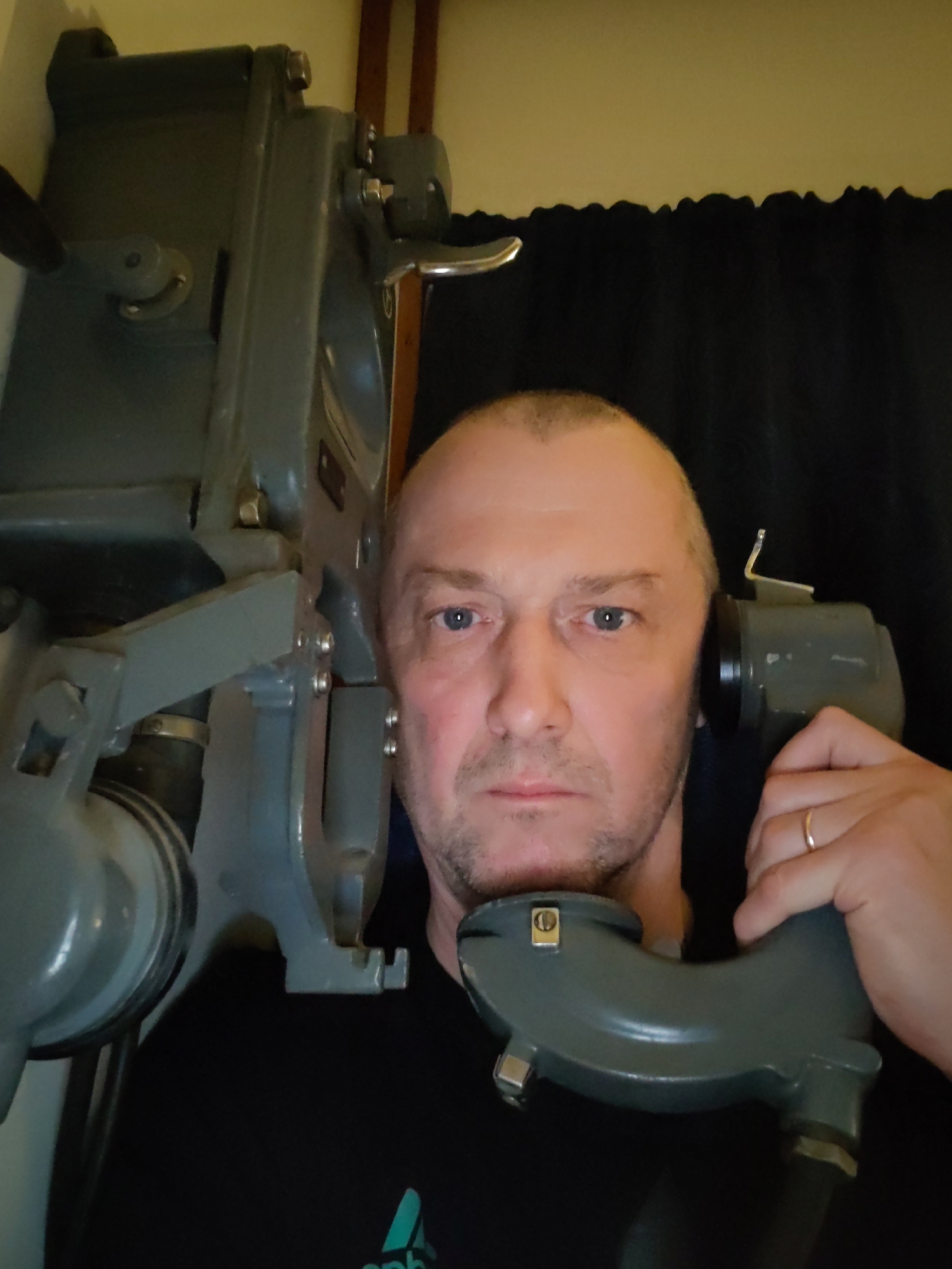 A man is posing with a vintage piece of equipment. The man has short, light brown hair, fair skin, and blue eyes. He is wearing a black shirt with a green logo visible at the bottom. He is holding a gray, antique-looking device to his ear. Another similar, larger device is beside him. The background is dark and nondescript, appearing to be a room with dark curtains. The location and city are unknown.