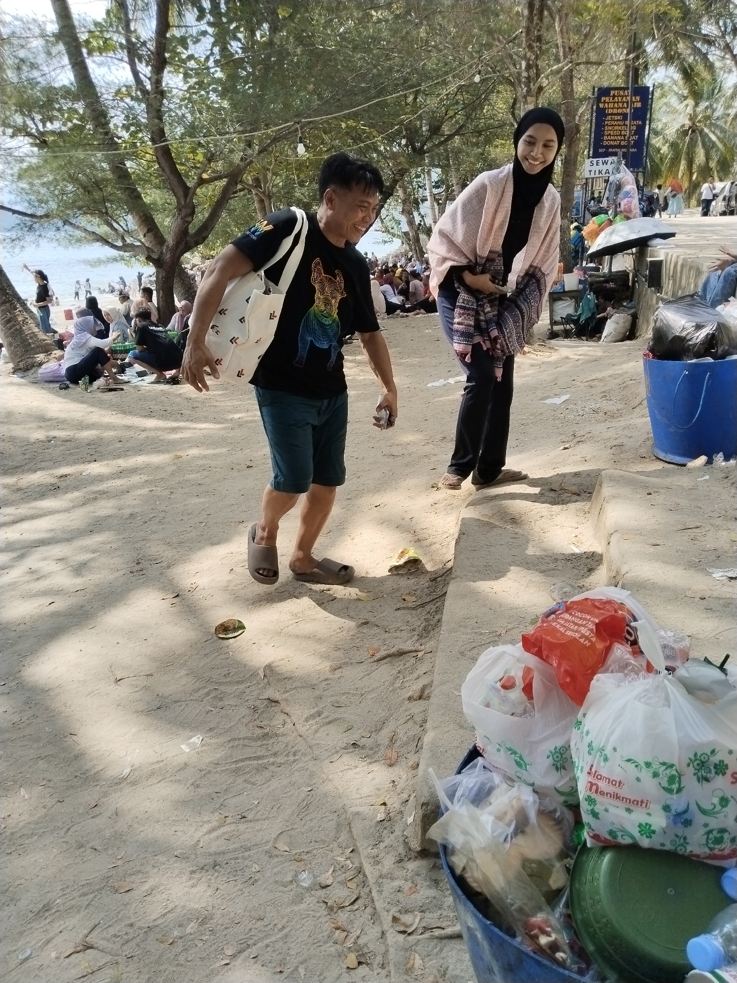 In Prigi, Indonesia, two individuals are visible on a sandy beach. A man in a black t-shirt and shorts, carrying a white tote bag, walks away from the camera, his body turned to the right. To his right, a woman wearing a black hijab and a pink cardigan over a dark top and pants smiles as she faces the camera. In the background, a crowd of people are gathered on the beach, some sitting and others standing. A blue trash bin overflowing with plastic bags and other refuse is situated to the right of the woman. A signpost with text in Indonesian is visible behind the trash bin, indicating services offered. Palm trees and other foliage frame the scene under a bright, sunny sky.