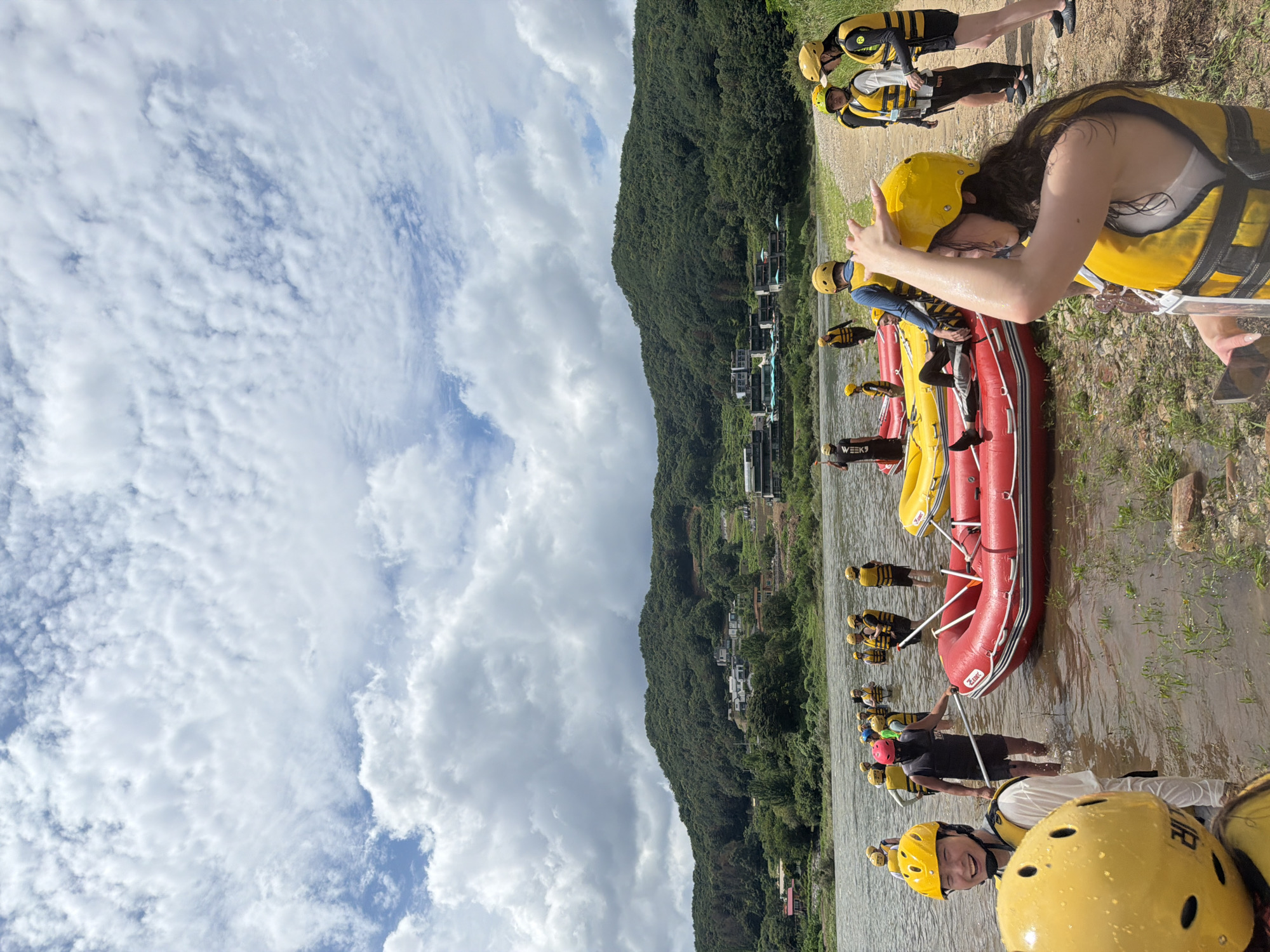 This is a photograph taken from the shore of a river in Seo-myeon, South Korea, under a partly cloudy sky. A group of people, predominantly wearing yellow life vests and helmets, are gathered by the water's edge and in the shallow river. Several inflatable rafts, red and yellow, are visible in the water and on the shore, indicating preparation for or participation in a water activity, likely rafting. In the background, lush green mountains rise, with some buildings visible on their slopes and a tree-lined riverbank. The people are engaged in various actions, such as wading, standing, and preparing the rafts. Some individuals are looking towards the camera, while others are interacting with each other or the equipment. The overall scene suggests an outdoor recreational setting on a summer day.