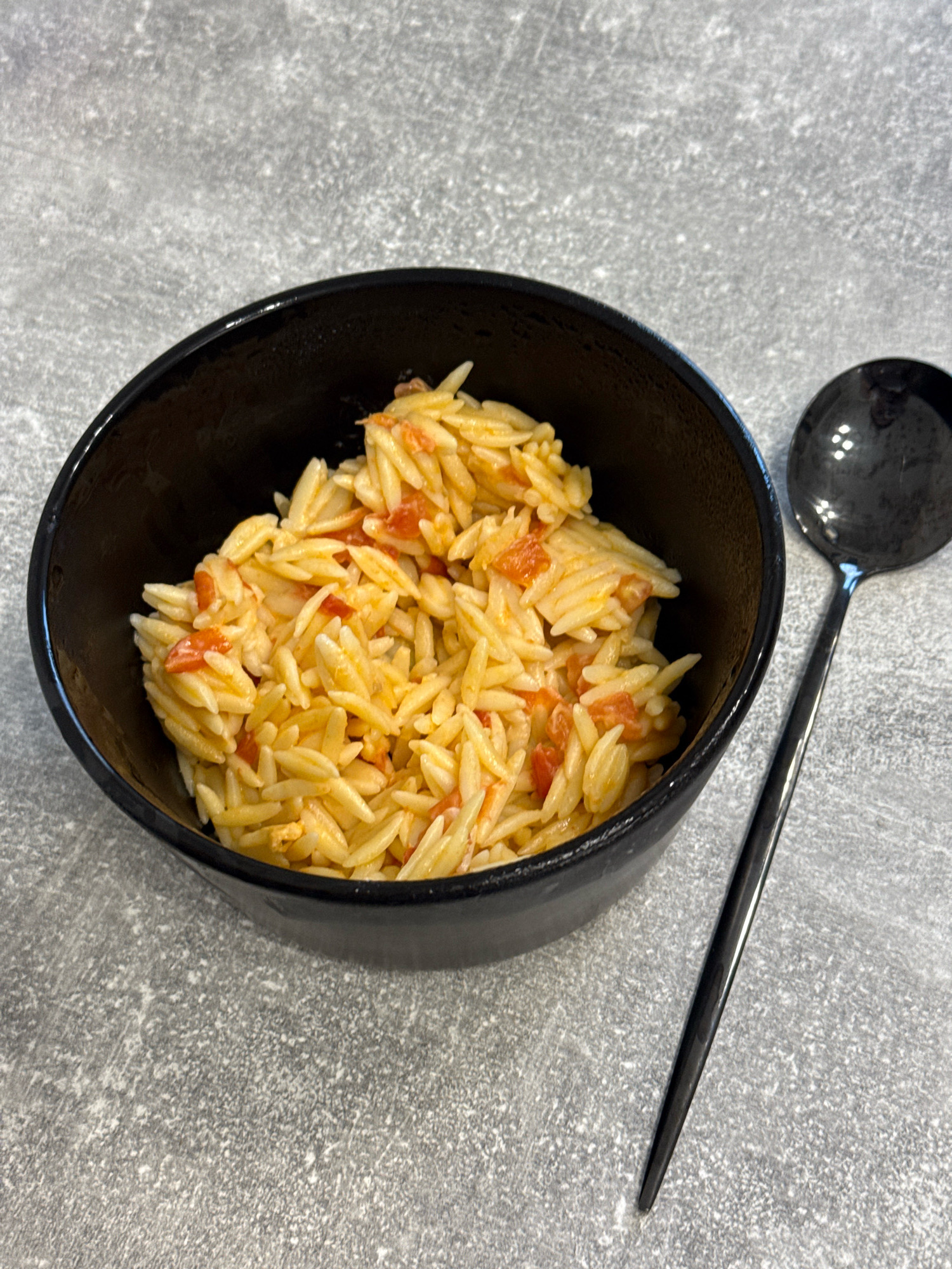 A black bowl filled with orzo pasta mixed with diced tomatoes sits on a textured gray surface. A black spoon with a rounded head rests to the right of the bowl, angled downwards. The image is a close-up shot, focusing on the food and spoon. There are no people visible, and the background is a uniform, speckled gray surface.