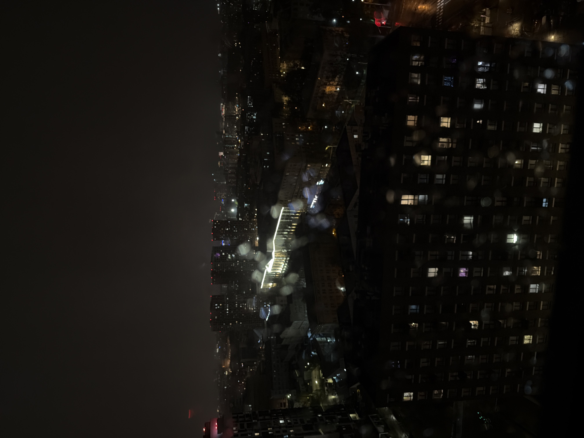 The image shows an aerial view of a city at night, possibly during or after rainfall, as evidenced by droplets on the window or lens. The lighting is dim, punctuated by city lights and street lights appearing as scattered points of brightness. On the right side, a tall building with illuminated windows dominates the scene, its repetitive pattern of lights giving it a structured appearance. To the left of this building stretches the cityscape, merging into a darkened area. A bright line of lights appears to be a major thoroughfare or a lit structure, cutting through the cityscape. The overall atmosphere is one of urban density and nighttime tranquility.
