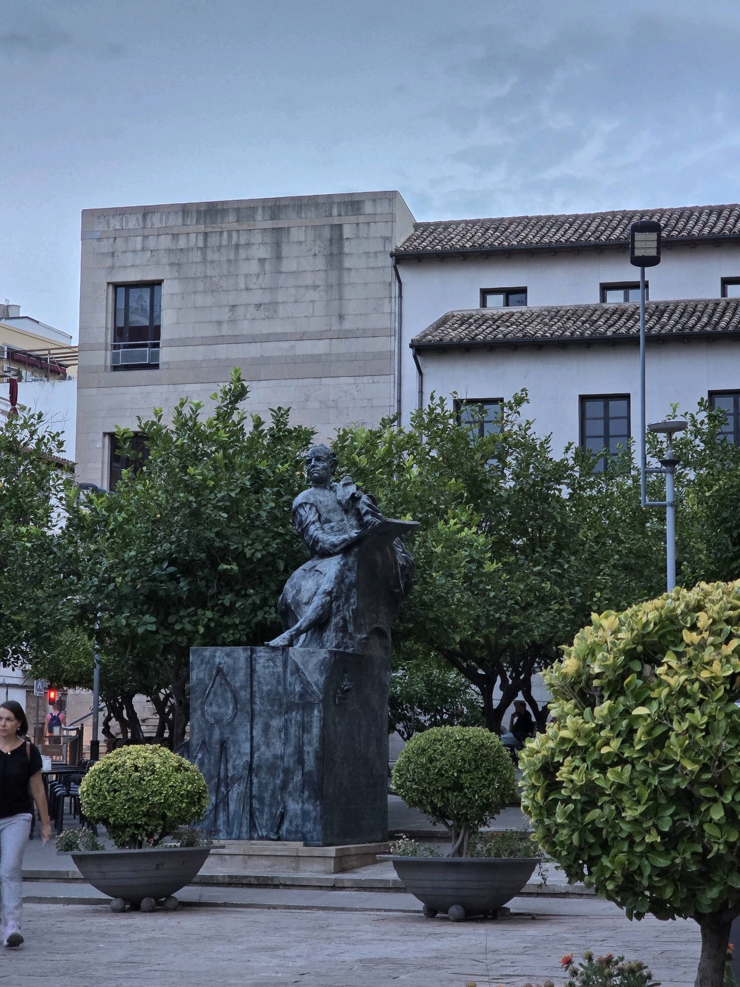 The image shows a bronze statue of a seated figure holding a book, likely a literary or historical figure, in a public square in Jaén, Spain. The statue is positioned in front of a building with a tiled roof and a pale facade, possibly an older structure, and a more modern, lighter-colored building with a stone exterior. Trees, some with lush green foliage and others appearing to be citrus trees, surround the statue. In the foreground, a woman with dark hair, wearing a black top and striped pants, walks across a paved area. Several decorative potted plants, one a spherical topiary and another a larger, fuller shrub, are visible in front of the statue. The sky is overcast, suggesting late afternoon or early evening light. A street light on a pole is visible to the right of the buildings.
