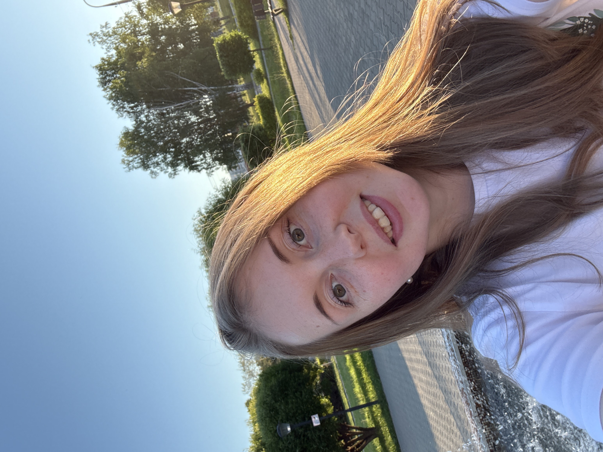 A young woman with long, blonde hair and a fair complexion is taking a selfie in a park. She is wearing a white t-shirt and a pearl earring. The sun is shining brightly, casting a warm glow on her face and hair. In the background, there are trees, bushes, a paved walkway, and a lamppost. The image is taken outdoors during the day.