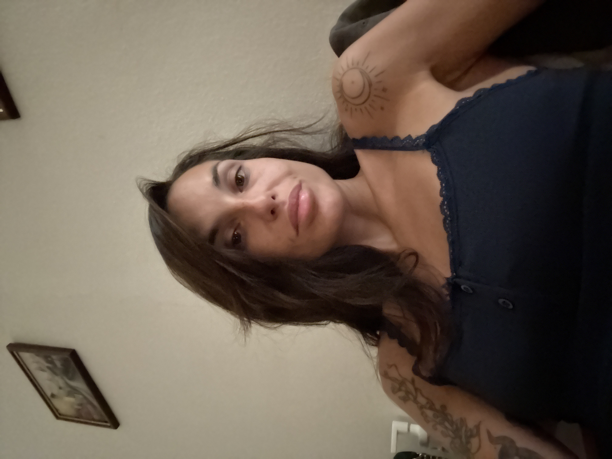 A medium close-up frontal portrait of a woman with dark brown hair and brown eyes is captured against a neutral-toned wall. She is wearing a dark blue tank top with lace trim and two dark buttons. She has tattoos on her left shoulder and arm, with a circular tattoo on her right shoulder featuring a crescent moon and sun. A framed picture is visible on the wall in the upper left corner. The lighting is dim, casting a shadow on the right side of her face.