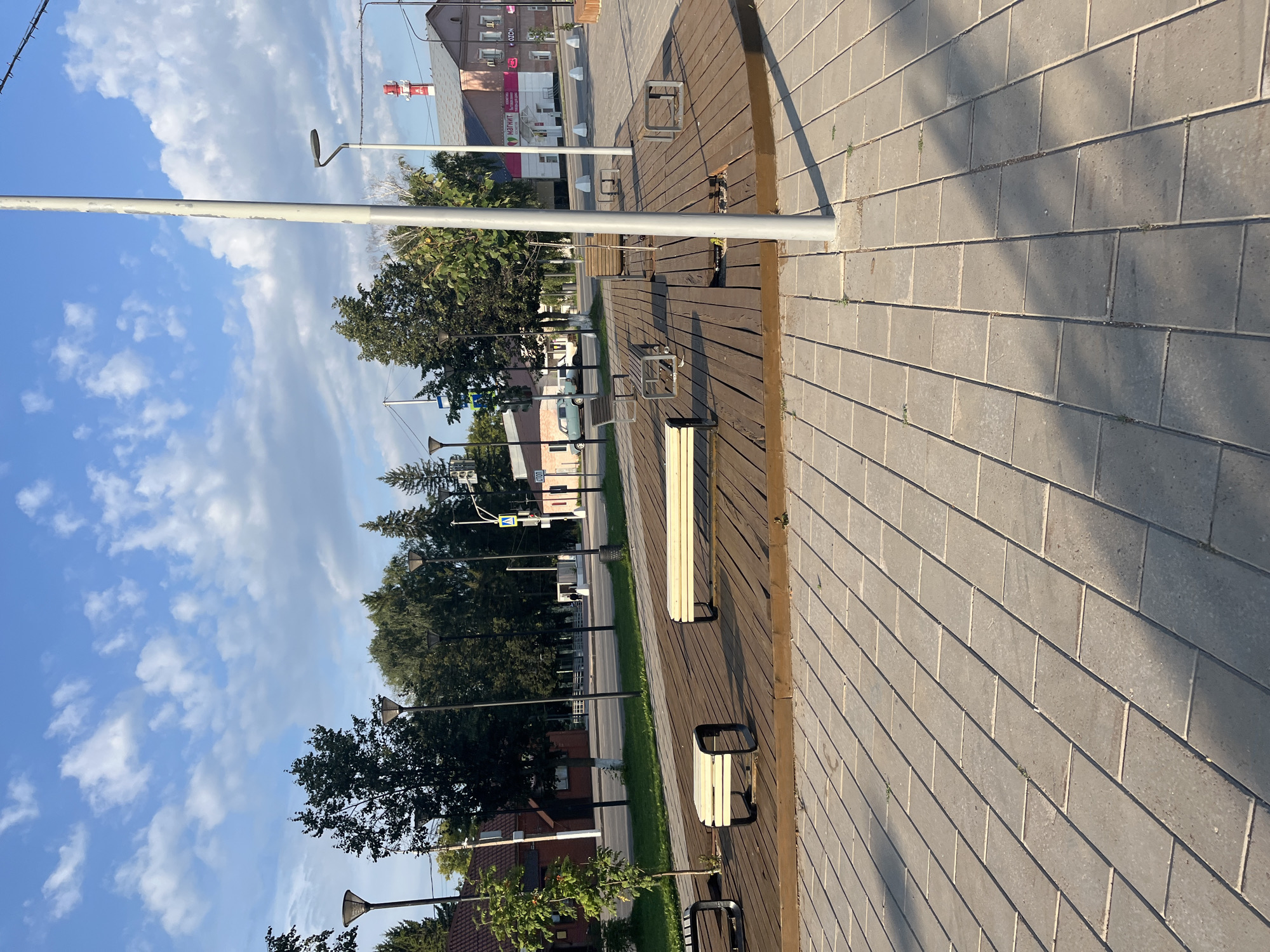 This image depicts a public space in Novye Turbasly, Russia, featuring a paved area with gray rectangular tiles in the foreground and a wooden deck with benches in the midground. Several modern streetlights are visible throughout the scene, as well as trees and buildings in the background. A tall, gray lamppost stands prominently in the center of the frame, casting a shadow on the pavement. The sky is blue with scattered white clouds. Buildings in the background include a pink and white structure with a red and white smokestack, possibly an industrial building or factory. A sign with Russian text is visible on one of the buildings. The scene appears to be set in the daytime under clear skies.