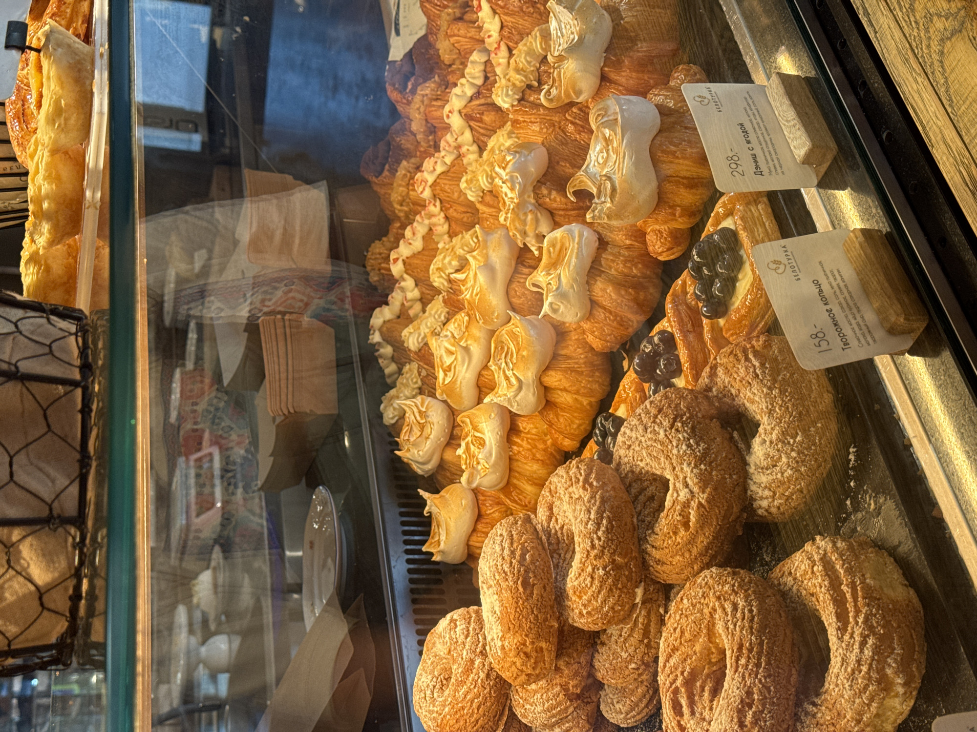 This media file shows a close-up, slightly angled view of a display case filled with pastries. The pastries appear to be in a bakery, with a glass barrier reflecting some of the surroundings. The primary subjects are a variety of baked goods, including croissants topped with white meringue, and ring-shaped pastries dusted with powdered sugar, some of which are topped with dark berries. There are also some elongated, flaky pastries visible.
Two price tags with Cyrillic text are prominently displayed. One tag reads "298.- Дэниш с ягодой" (Danish with berries) and the other reads "158.- Творожное кольцо" (Tvorozhnoe koltso - which translates to cottage cheese ring). The text also includes bakery branding "БЕЛОТУРКА". The lighting suggests it could be daytime, possibly indoors with artificial light complementing natural light. The scene is static, focusing solely on the food display. The overall impression is one of abundance and a tempting selection of sweet treats, likely in a bakery located in Izhevsk, Russia, as indicated by the Cyrillic text and location context. The pastries are arranged in a visually appealing manner, suggesting a commercial setting.