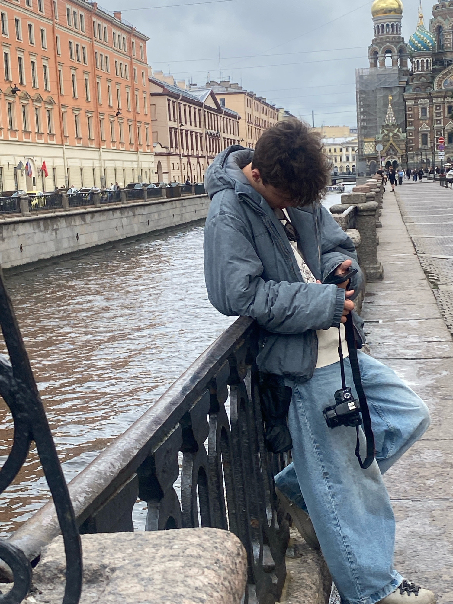 A young person with curly brown hair, wearing a grey puffer jacket, a white t-shirt, and baggy light-wash jeans, leans against a black metal railing overlooking a canal. They are looking down at their phone, holding it in their right hand, while a black camera with a strap hangs around their neck. The canal water is a murky brown, reflecting the overcast sky.
In the background, on the left, is a row of pink buildings with many windows. On the right, the iconic Church of the Savior on Spilled Blood, with its ornate domes and multi-colored facades, is visible. The ground is paved with cobblestones, and other pedestrians can be seen walking along the embankment. The overall scene depicts a cloudy day in Saint Petersburg, Russia, likely near a popular tourist area.