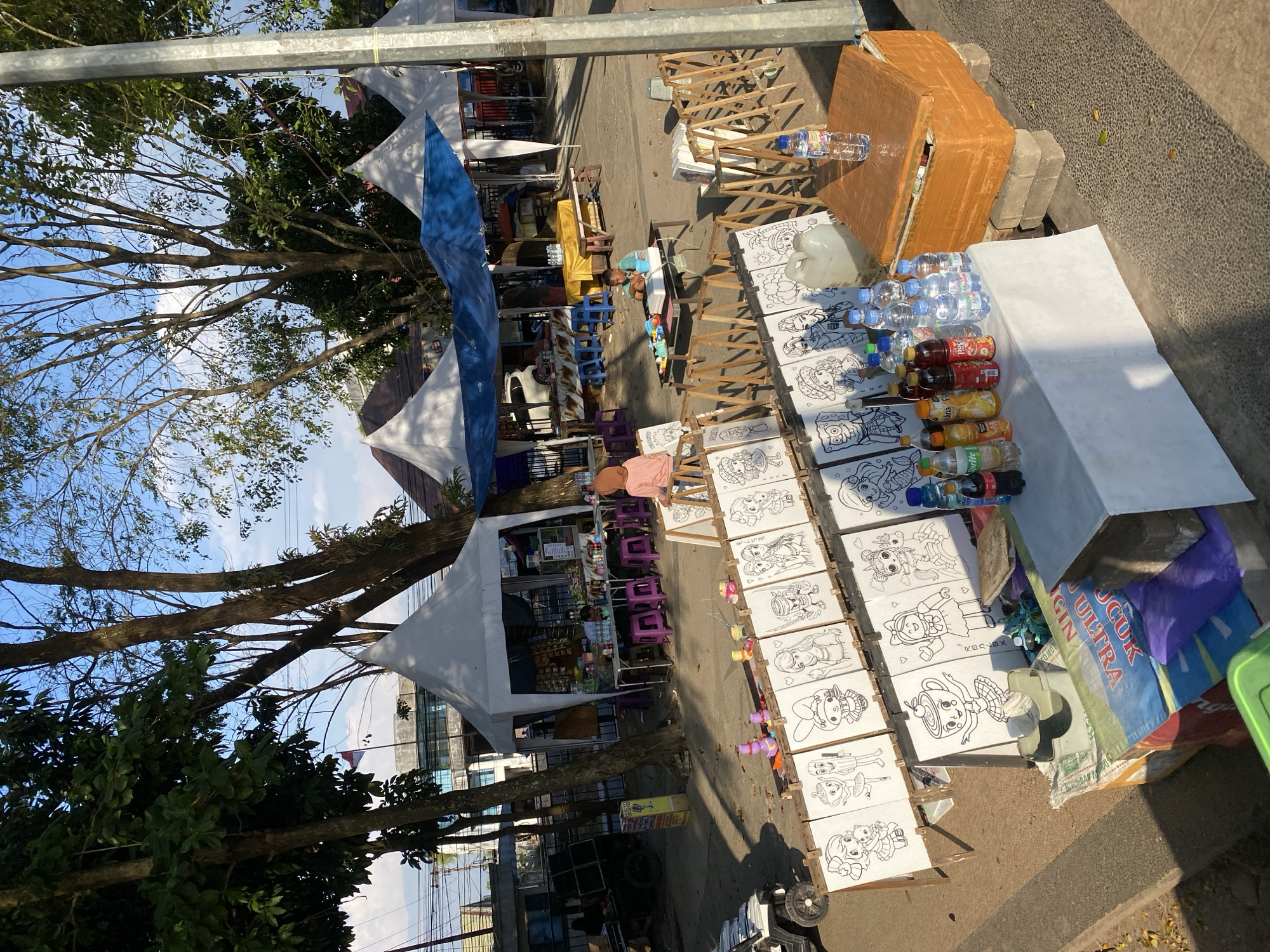 The image depicts an outdoor market scene in Banjarmasin, Indonesia. The foreground is occupied by a display of children's coloring pages featuring various cartoon characters, arranged on easels. To the right of the coloring pages, a white table is set up with an assortment of bottled beverages, including water, soda, and juice. An orange cooler box sits on top of stacked bricks beside the table. In the background, several white and blue tents are set up, suggesting food or merchandise stalls. A woman wearing a hijab and a pink shirt is seated on a stool near one of the tents. The environment appears to be a public area with trees providing shade, and the ground is a mix of paved and dirt surfaces. The overall atmosphere suggests a casual market or event taking place outdoors during daylight hours.