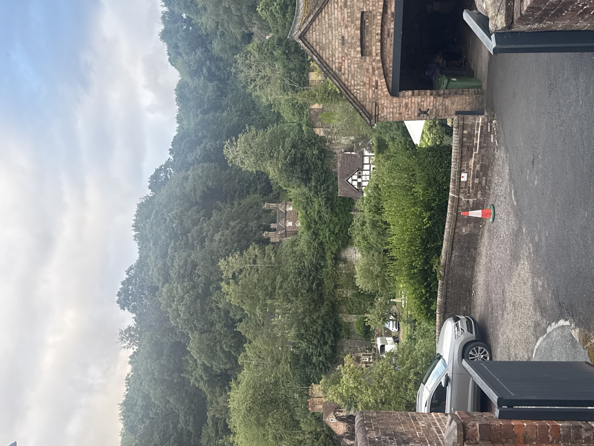 This image depicts a scene in Ironbridge, United Kingdom. The perspective is from a raised vantage point, looking down towards a wooded hillside and a street. In the foreground, a brick building with an arched opening, possibly a covered entryway or garage, is visible. A gray SUV is parked on the asphalt road in front of this building. Further down the road, a white traffic cone is placed on a brick wall.
The background features a densely wooded hillside with various buildings nestled amongst the trees. One building has a distinctive black and white timber-framed facade. The sky is overcast, suggesting it is daytime but potentially a cloudy or damp day. The overall atmosphere is somewhat subdued due to the weather and the green, natural surroundings. There is no discernible activity or event taking place; it appears to be a still, observational shot of the location. No visible text is present in the image.