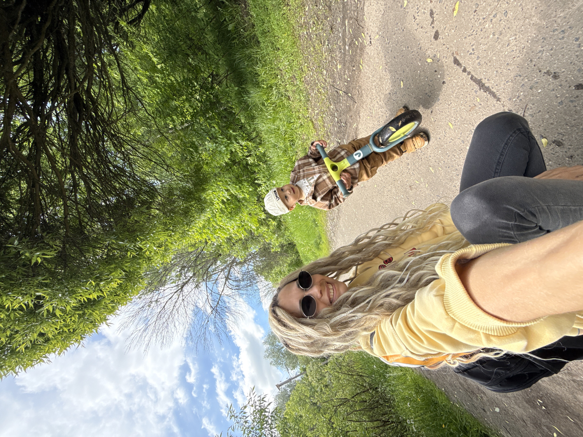 A sunny outdoor photograph depicts a woman and child in Rusanovo, Russia. The woman, wearing a yellow sweater, black jeans, and round sunglasses, smiles toward the camera. She has long blonde hair. Next to her, a young boy wearing a plaid shirt and a white cap sits on a small yellow and blue balance bike. They are on a paved path next to green grass and trees. Above, blue sky and clouds can be seen through the tree branches.