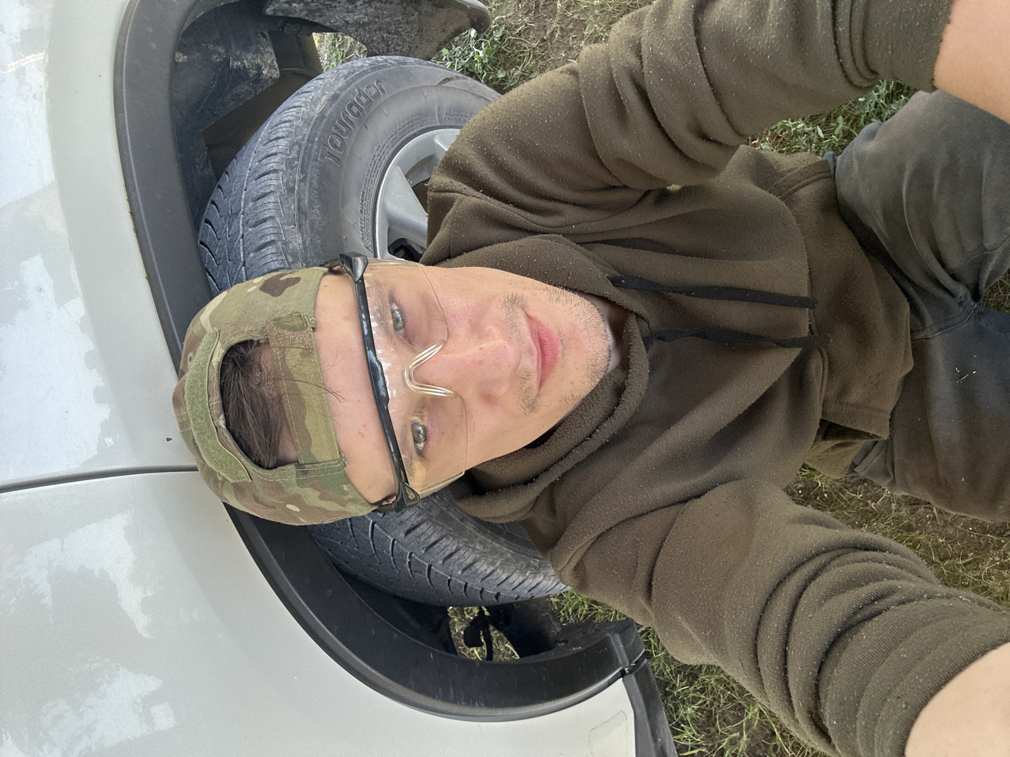 A person is shown in a selfie, crouching next to the wheel of a white vehicle. The person is wearing a camouflage baseball cap, clear protective glasses, and a dark green hooded sweatshirt. The vehicle appears to be an SUV, and its tire is visible, with the brand name "Tourador" discernible. The ground is covered with grass and dirt. The image was captured outdoors, likely in a rural or suburban setting. The exact location within the city of Makiv, Ukraine, cannot be determined from the image alone, as there are no identifiable landmarks or signs.