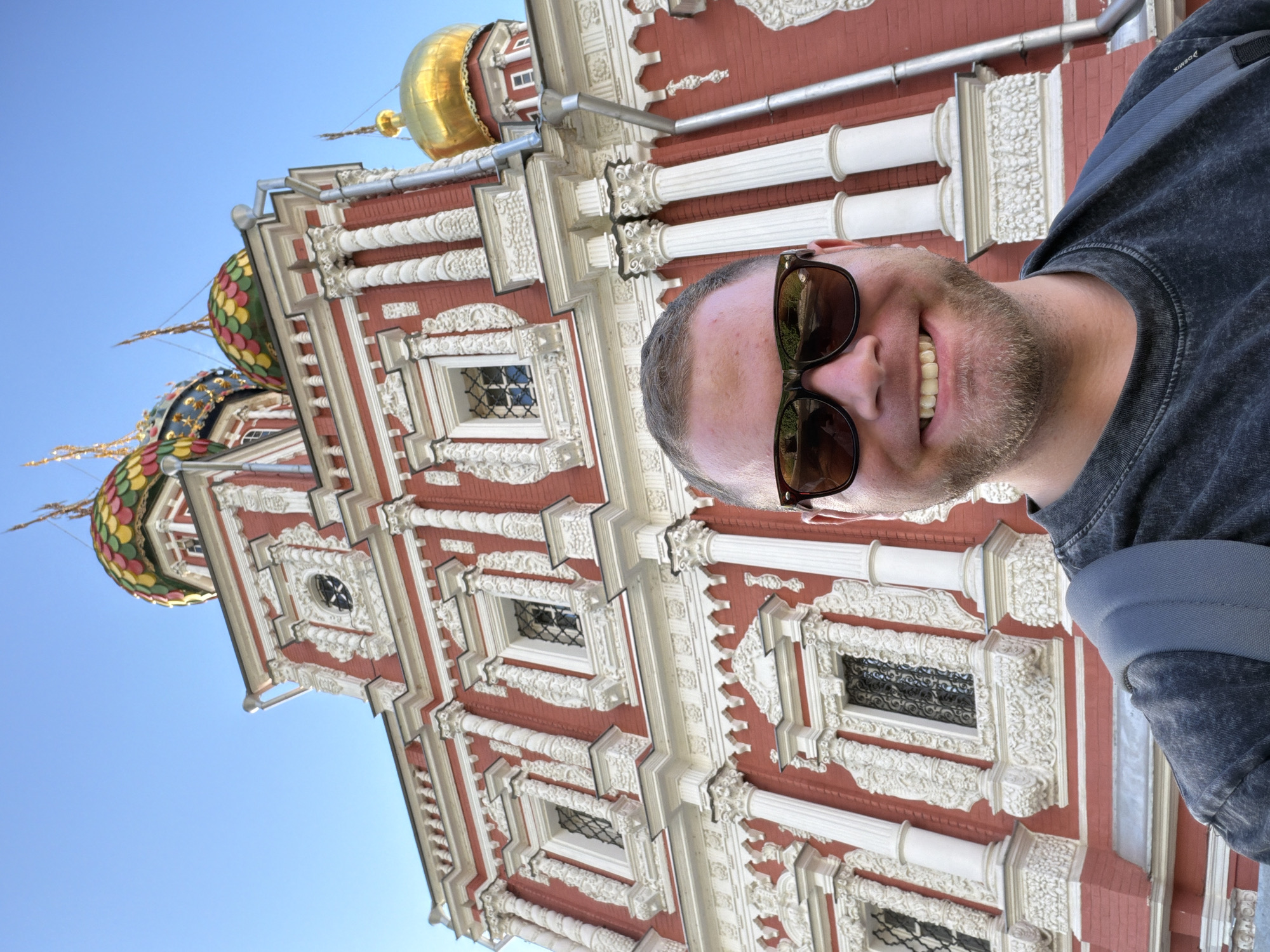A man with short brown hair and a beard, wearing sunglasses and a dark t-shirt, smiles broadly for a selfie. He is positioned in the foreground, with a portion of his shoulder and backpack visible. Behind him, a vibrantly decorated building with red walls and intricate white ornamentation dominates the frame. The building features multiple ornate domes, one gilded and others in colorful mosaic patterns of green, yellow, red, and blue. The architectural style suggests a historical or religious building, likely a church or cathedral. The sky is clear and bright blue, indicating a sunny day. The image is oriented vertically, with the top of the building appearing at the bottom of the frame and the man's head at the top. The man's expression is joyful and relaxed. The scene appears to be outdoors in a location with significant architecture, consistent with Nizhny Novgorod, Russia.
