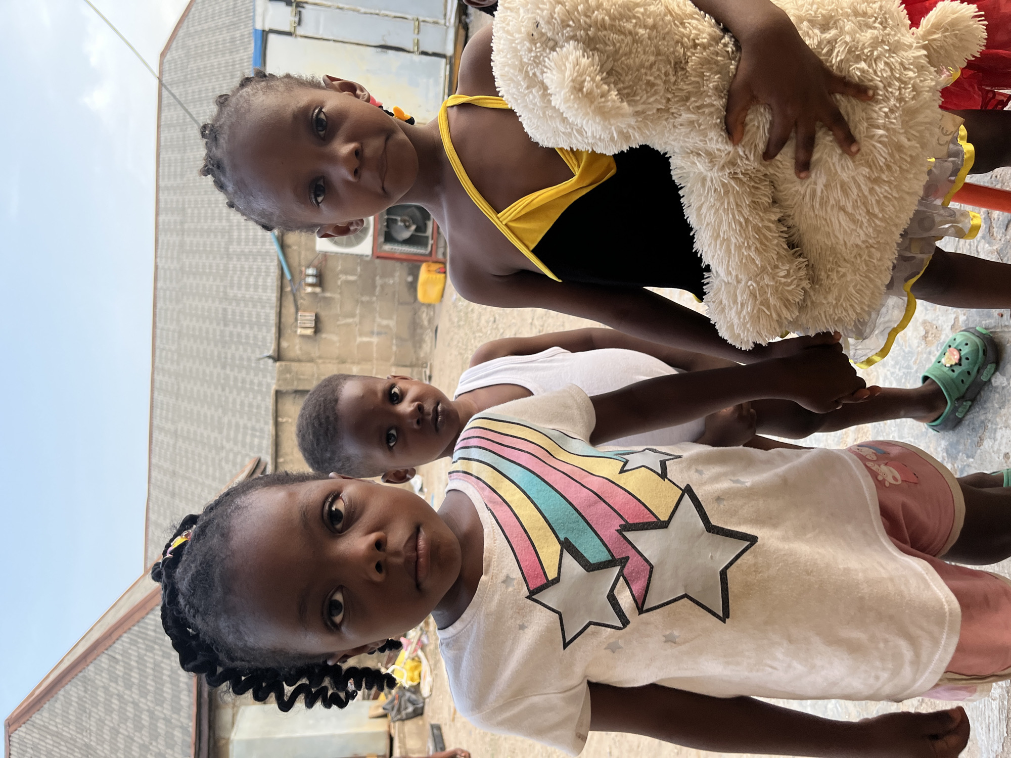 Three young African children are posing for a photo outdoors. One girl wears a white t-shirt with rainbow stripes and star embellishments. Another wears a black top with yellow straps and holds a light-colored stuffed animal. The third child wears a white tank top. The background includes a light blue sky and buildings constructed of gray blocks. The location is Iddah, Nigeria.