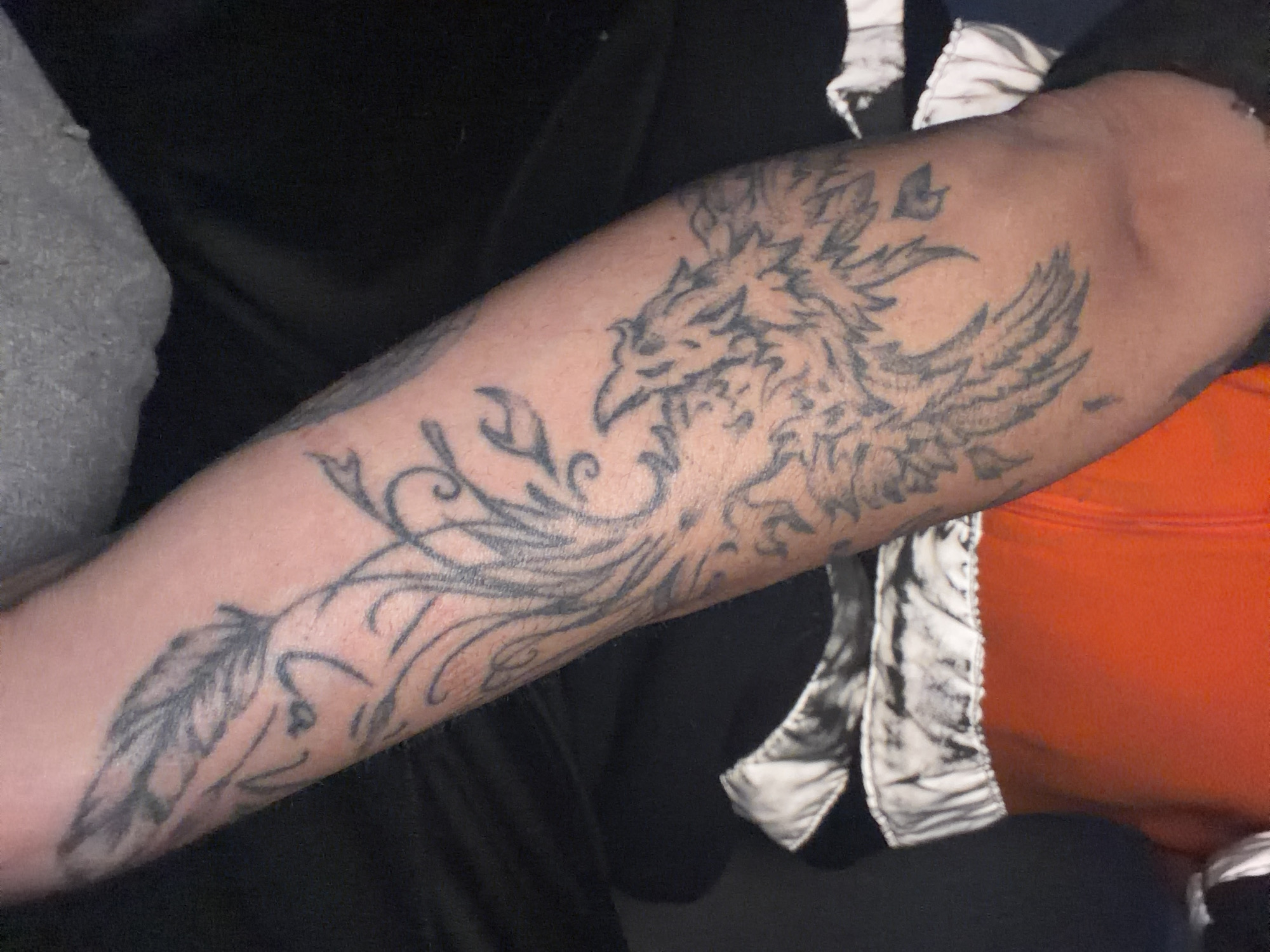 The image shows a person's forearm with a large tattoo of a phoenix with feathers and swirling patterns. The tattoo is in black ink. The person is wearing a gray shirt on the left and an orange garment with a white and black patterned item on the right. The background is dark and blurry. There is no information indicating this image was taken in Sant Martí Sarroca, Spain.