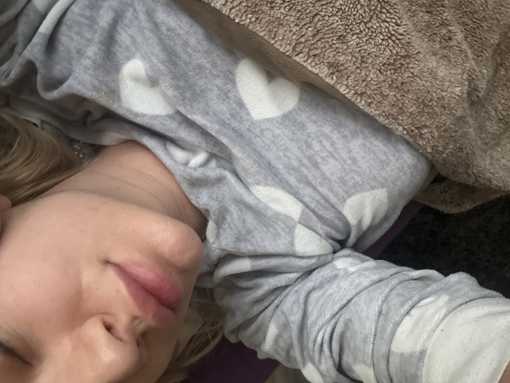 The image presents a close-up, angled view of a person resting comfortably indoors, likely in Sofia, Bulgaria. The subject appears to be a woman, with fair skin and visible blonde hair peeking from behind her head. Her eyes are closed, and her facial expression suggests peaceful sleep or deep relaxation, with a slight, natural parting of her lips.
She is wearing a soft, gray long-sleeved top or covered by a blanket, characterized by a pattern of white hearts. The fabric shows gentle wrinkles, indicating movement and comfort. Part of her arm, covered by the same heart-patterned material, is visible in the lower right. A brown, textured, fluffy blanket or fabric is also visible in the upper right portion of the frame, providing an additional layer of warmth and coziness. The overall setting implies an intimate and comfortable environment, such as a bed or sofa in a home. The lighting is soft and diffuse, contributing to the tranquil atmosphere. There is no visible text in the image.