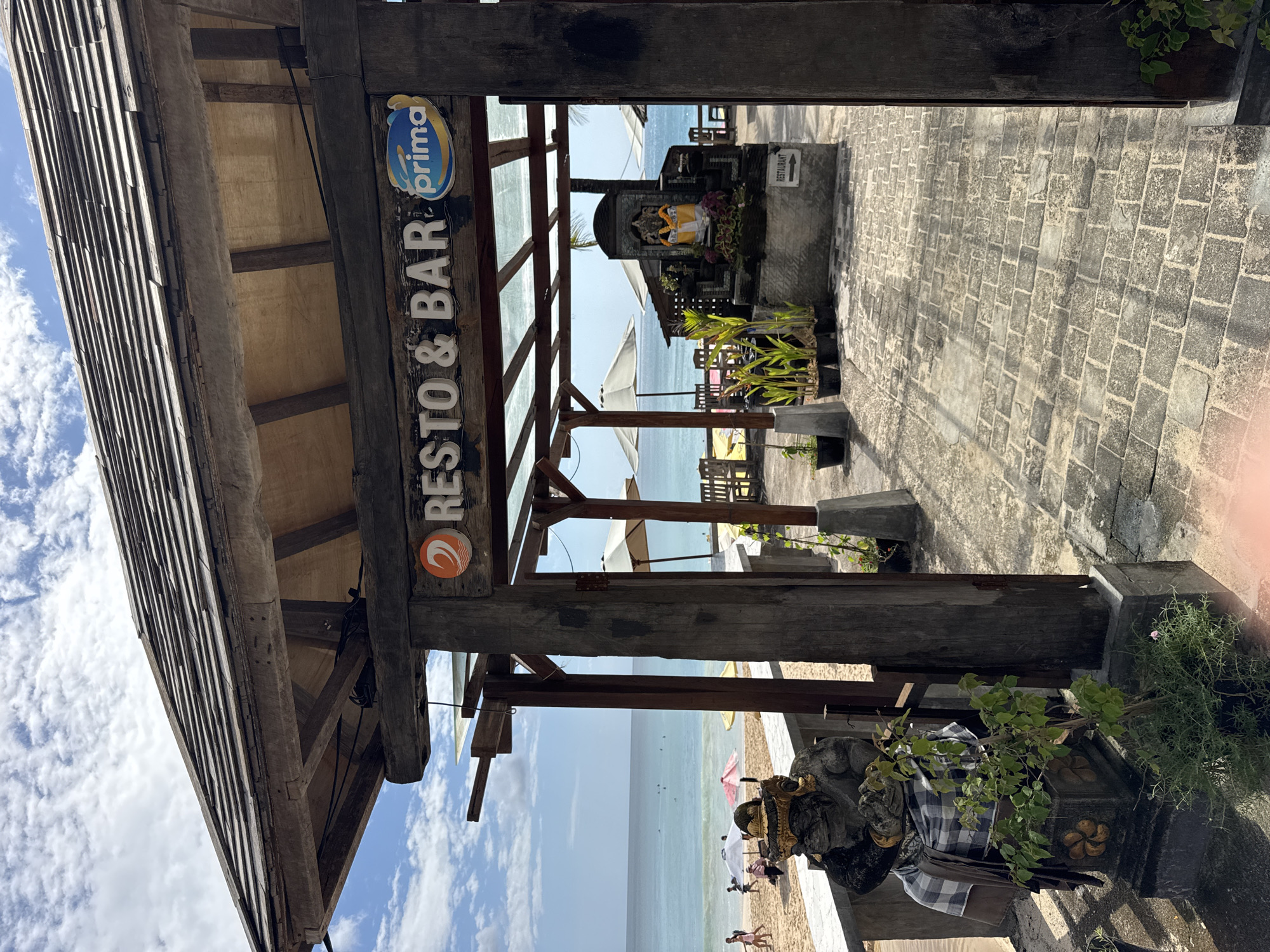 The image captures the entrance to a "RESTO & BAR" in Kuta Selatan, Indonesia. The entrance is framed by a wooden structure with a sign displaying the name and a "Prima" logo. Beyond the entrance, a stone-paved walkway leads towards the beach. A Balinese shrine is visible along the walkway, adorned with offerings. Beach umbrellas and chairs suggest a tourist-oriented establishment. People are present on the beach and in the water. The sky is blue with scattered clouds. A sculpture wrapped in a checkered cloth stands to the right of the entrance.
