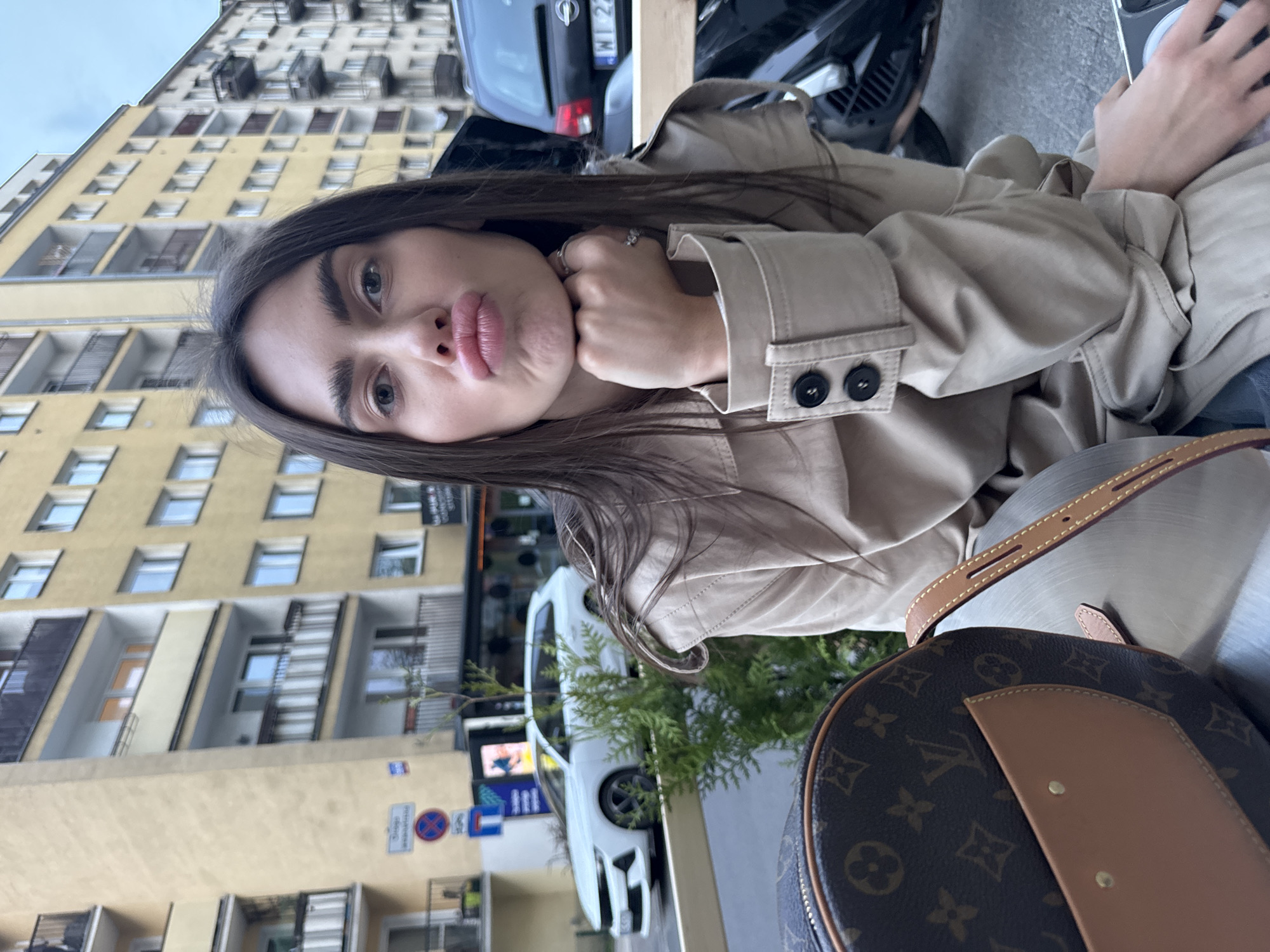 A young woman is in Warsaw, Poland. She is seated, wearing a beige trench coat, and posing for a selfie with pursed lips. A Louis Vuitton bag is near her. Behind her is a street with visible traffic signs, a white car, and a parked black car with a Polish license plate that appears to read "WI 22". A tall yellow building stands in the background.