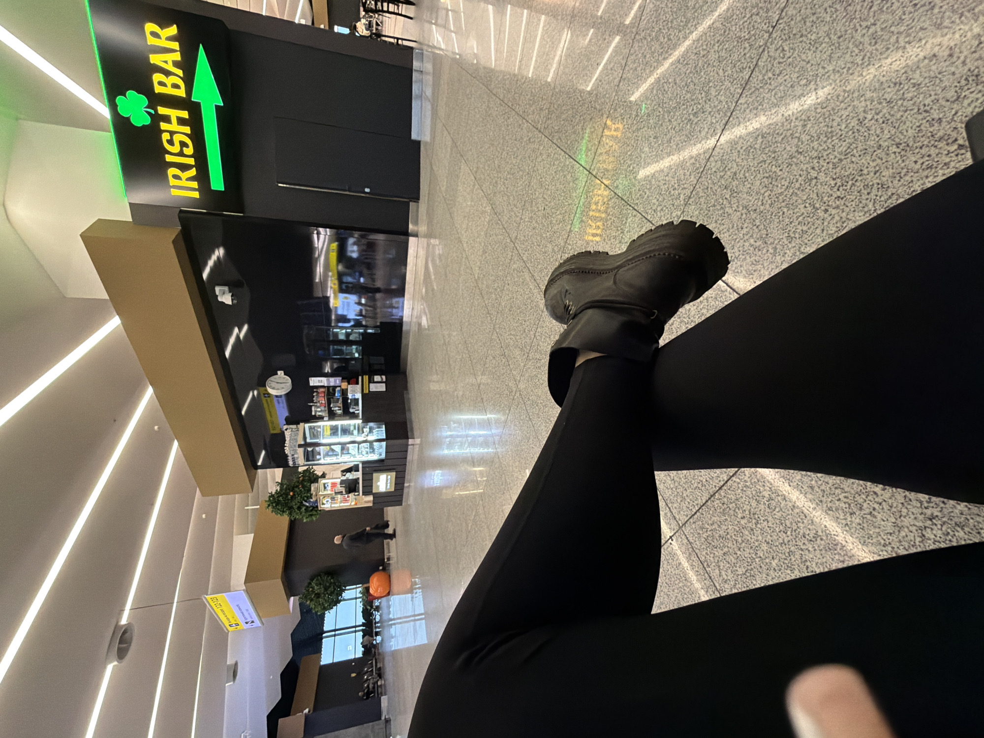 The image captures a view from a seated position in an airport, presumably in Khimki, Russia, based on the possible location context. The individual is wearing black leggings and a black boot on their left foot, which is crossed over the right leg. In the background, there's an "IRISH BAR" sign with a green arrow pointing towards the bar's entrance. A person in a dark coat walks in the distance, and there is a row of shops with glass display cases. The environment suggests a typical airport terminal with grey-tiled flooring and bright lighting. The image appears to be taken indoors with the camera focused on the seated individual and their legs.