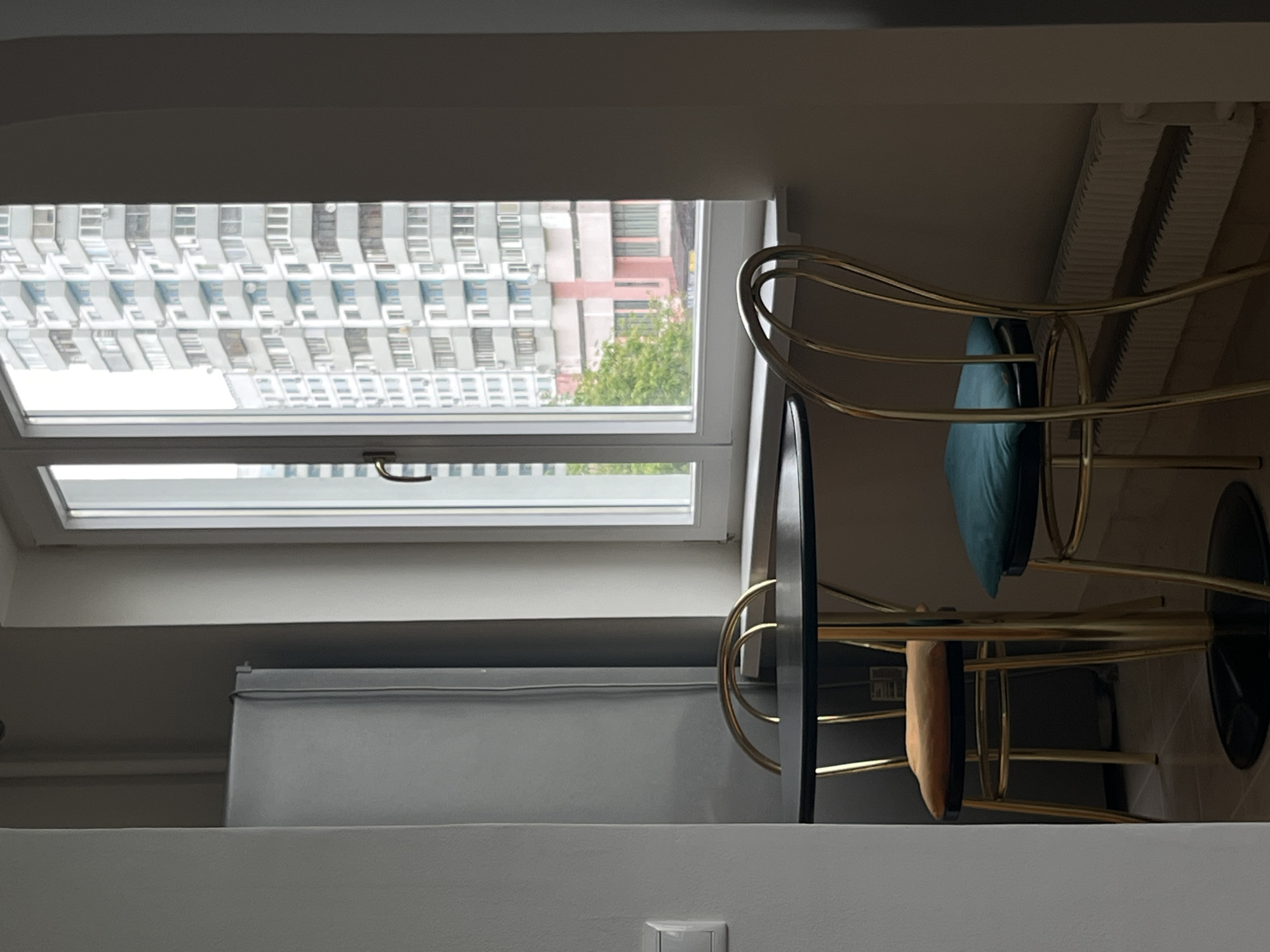 The image shows an interior view with a window overlooking a dense urban landscape. The architecture outside the window suggests a high-rise residential area, likely in a large city. Based on the architecture, this could be Moscow, Russia. The window has a horizontal handle and is multi-paned.
Inside, a contemporary metallic gold bar stool with two cushions is visible. The scene is well-lit but with strong contrasts between light and shadow. The window appears to be a skylight. A white object on the wall is visible, possibly an electrical outlet.