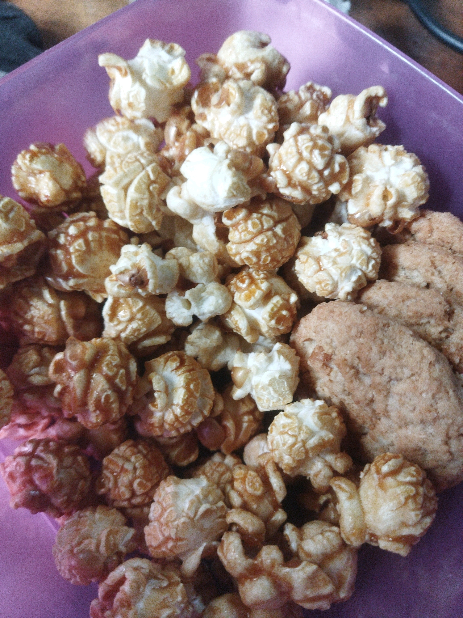 A close-up, overhead shot shows a purple plastic bowl overflowing with caramel popcorn and three round, tan-colored cookies. The popcorn pieces are irregular in shape, with some appearing to be coated in a shiny caramel glaze. The cookies are textured, with visible oats or grains. The scene is lit from above, casting soft shadows. The location is not specified in Tarai Bangun, Indonesia.