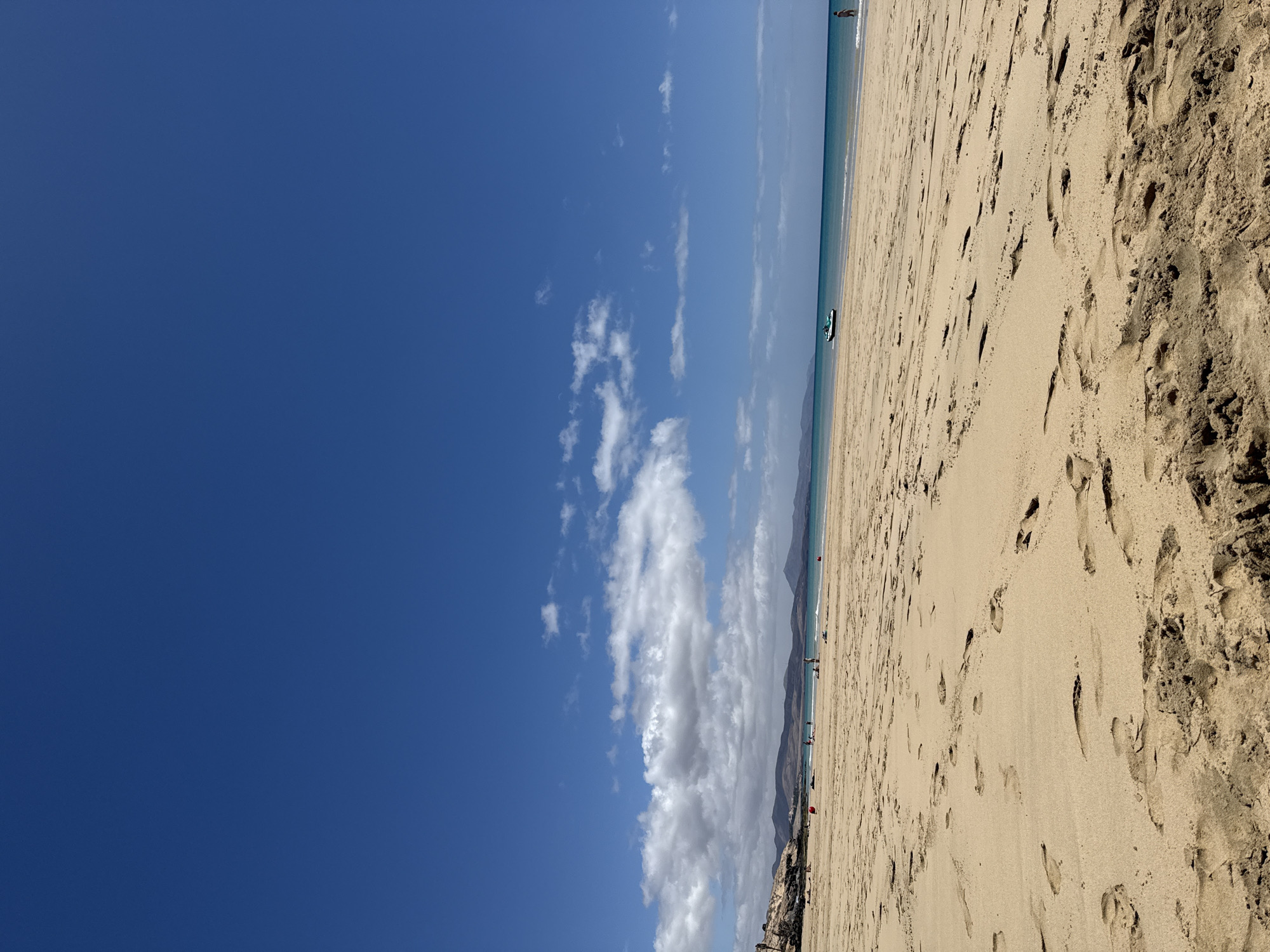 The image shows a beach in Pájara, Spain. The sand is a light brown color and covered in footprints. The sky is a clear blue with some white clouds. There is a body of water in the distance. There are a few people visible on the beach and in the water.