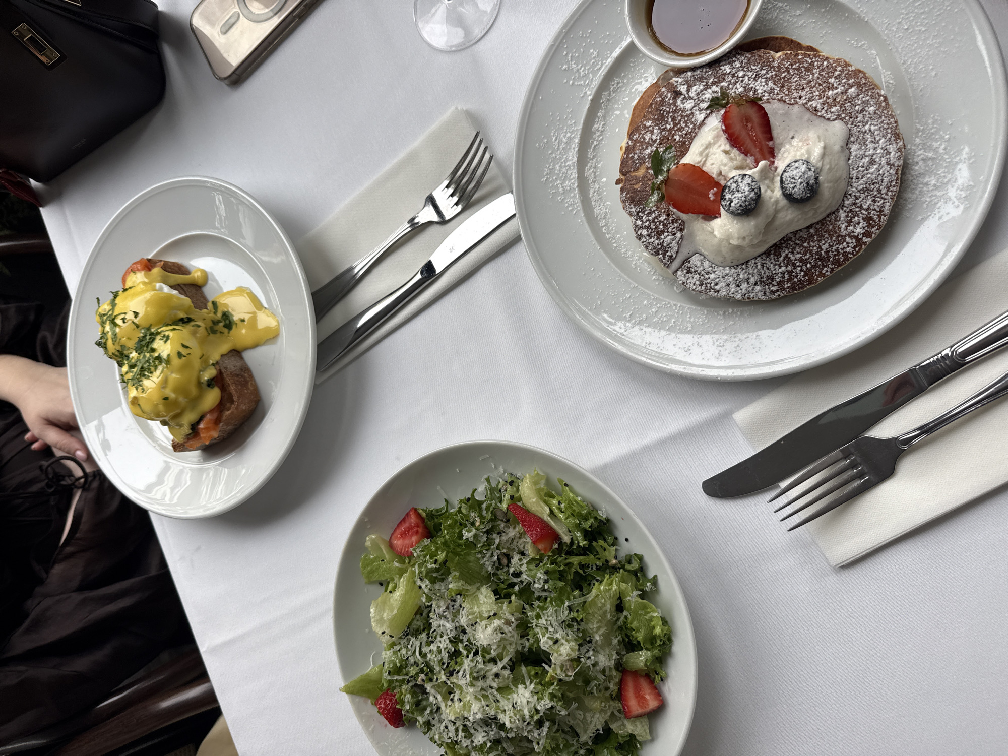 An overhead shot captures a table set for a brunch or breakfast meal, likely in Tel-Aviv, Israel. The table is draped with a clean white tablecloth, showcasing three distinct dishes and dining essentials.
In the top-left, a white plate holds two servings of Eggs Benedict. Each features a poached egg generously covered in bright yellow hollandaise sauce, garnished with finely chopped green herbs, possibly chives. These are served on toasted bread or buns, with hints of smoked salmon visible beneath one of the eggs. Beside this plate, a person's arm in a dark garment is partially visible, along with a black leather handbag with a gold clasp and a light-colored smartphone with a magnetic (MagSafe) attachment.
To the right, a large, round pancake is presented on a white plate. It is generously dusted with powdered sugar and topped with a dollop of white cream, fresh sliced strawberries, and whole blueberries. A small ramekin of maple syrup accompanies the pancake on the side.
At the bottom of the frame, a white bowl contains a fresh green salad. It consists of various lettuce leaves, interspersed with grated white cheese, scattered black seeds, and sliced fresh strawberries.
Silverware, including forks and knives, rests on folded white napkins beside the plates, indicating a prepared dining setup. A clear glass is partially visible near the pancake dish. The overall scene suggests a pleasant and varied meal, possibly enjoyed at a restaurant or cafe. No specific text is legibly visible on any items.