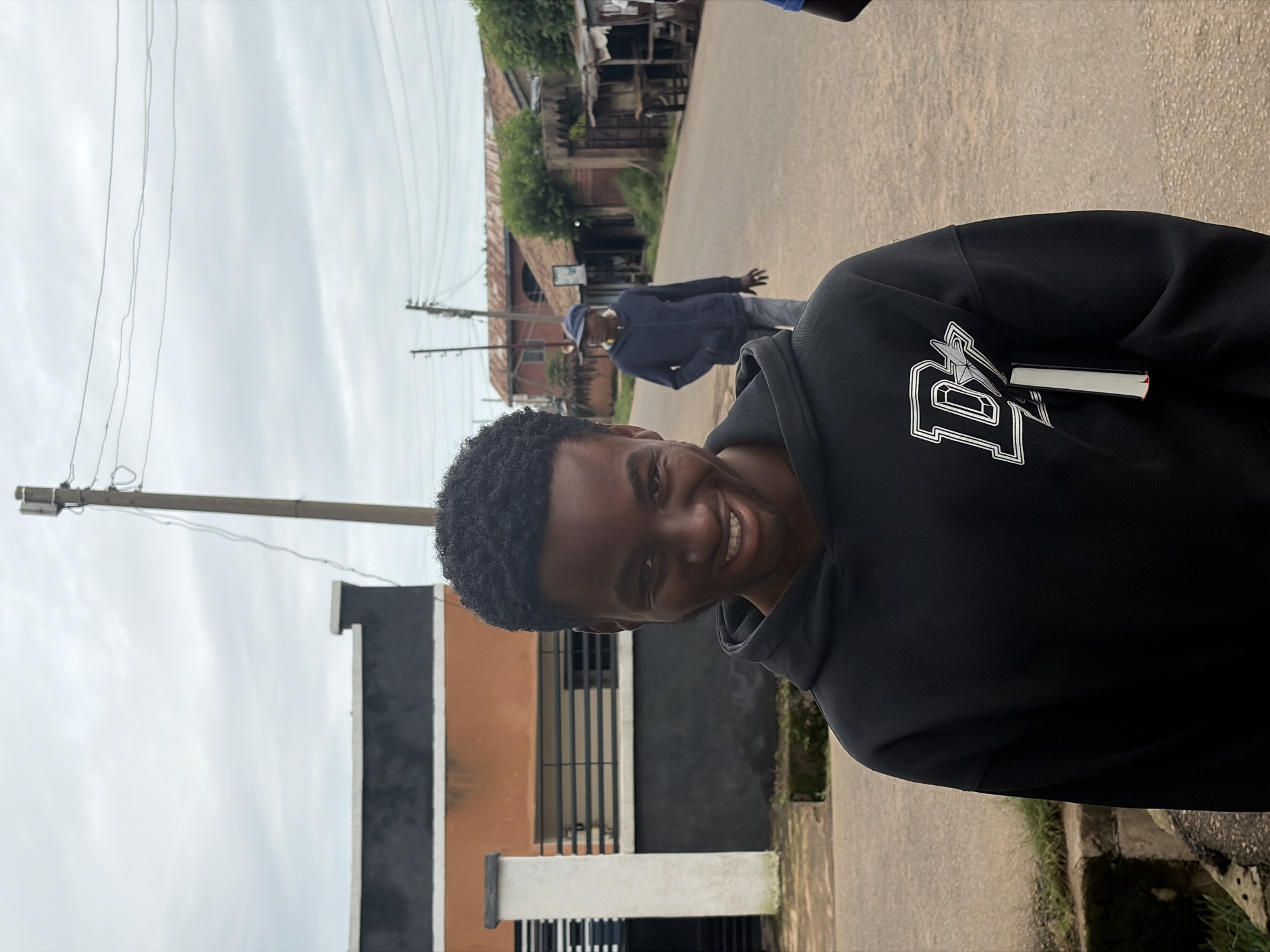 A young Black man with short, dark, curly hair and a wide smile is captured in a close-up, medium shot, facing the camera outdoors. He is wearing a black hooded sweatshirt with a white emblem featuring the letter "D" with a star inside, and a white object is tucked into the front pocket. To his right, a man in a dark blue hoodie and a blue cap walks away from the camera down a paved street. The background shows a street with buildings on either side, including a structure with orange and dark gray walls, and a telephone pole with wires. The sky is overcast. The scene is a residential street in Jos, Nigeria.