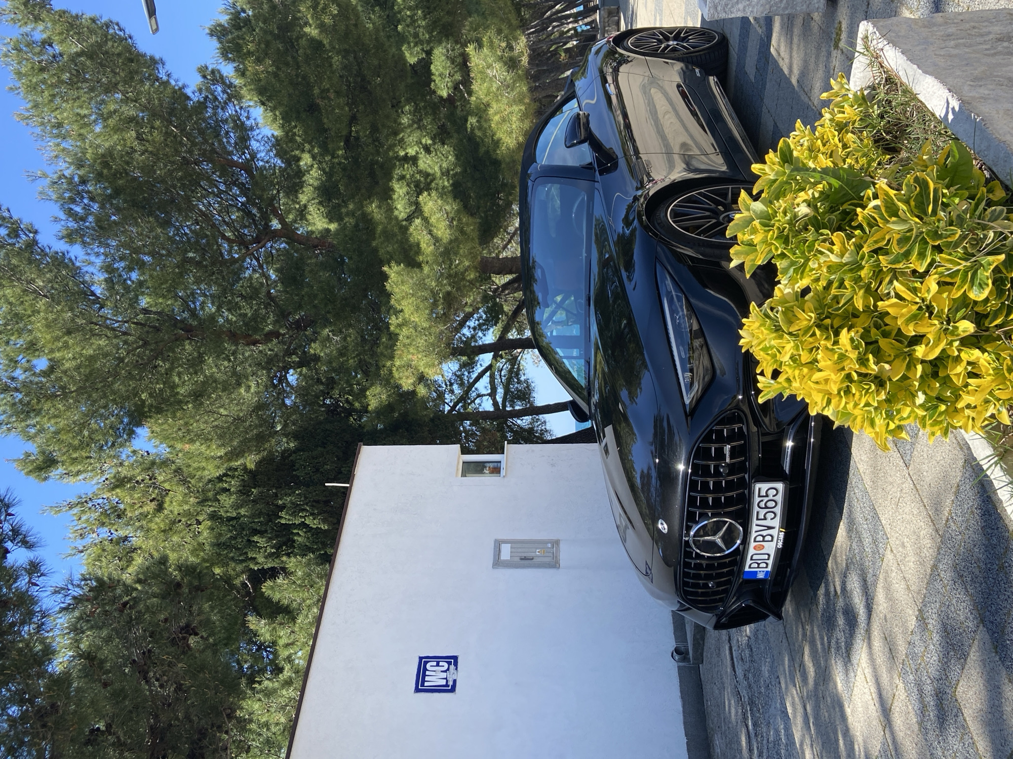 A black Mercedes-Benz, license plate "BD BV 565", is parked next to a small, white building with two small windows and a "WC" sign on the lower portion of the wall. The scene also includes vegetation; tall trees are behind the building, and a bush with yellow and green leaves is next to the car, with stone paving in the foreground. The photo was taken during the day. The city is Bar, Montenegro.