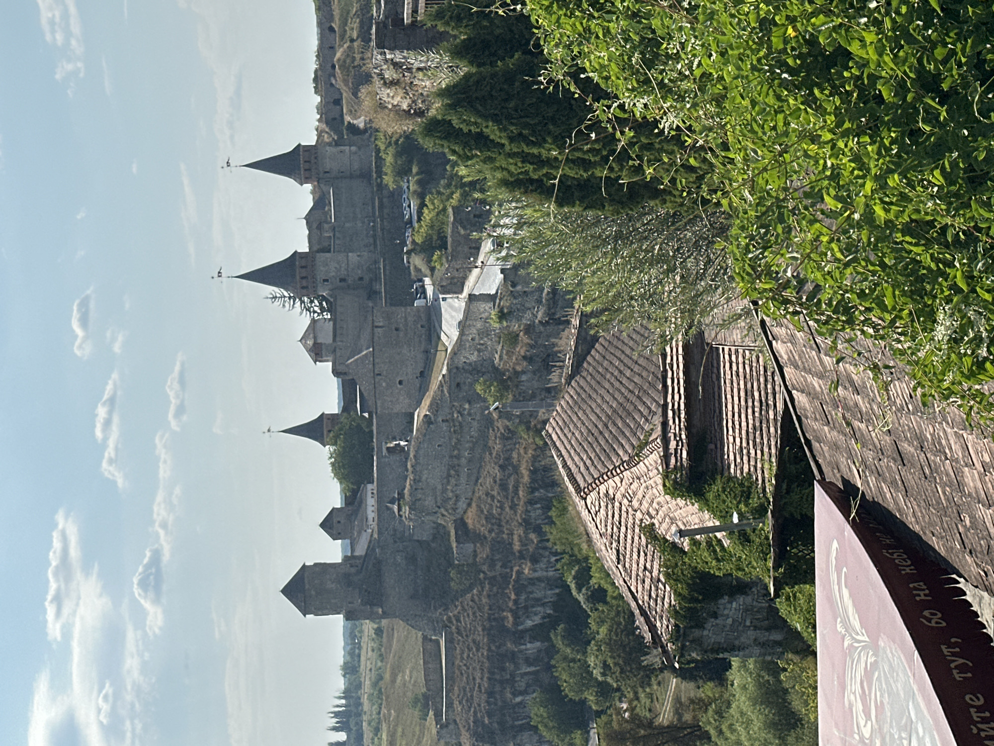 This is a photograph of the Kamianets-Podilskyi Castle in Kamianets-Podilskyi, Ukraine, taken on a bright, partly cloudy day. The castle, a large stone fortress with multiple towers and walls, sits atop a rocky hill. In the foreground, several tiled roofs of buildings are visible, partially obscured by lush green foliage. A road winds its way towards the castle, with a few parked cars near its entrance. The castle complex itself appears extensive, with a variety of architectural styles and stone construction. The surrounding landscape is hilly and green, with distant trees dotting the horizon. There are no people visible in the image. The overall impression is of a historical landmark set within a natural environment, under a clear blue sky with scattered white clouds.