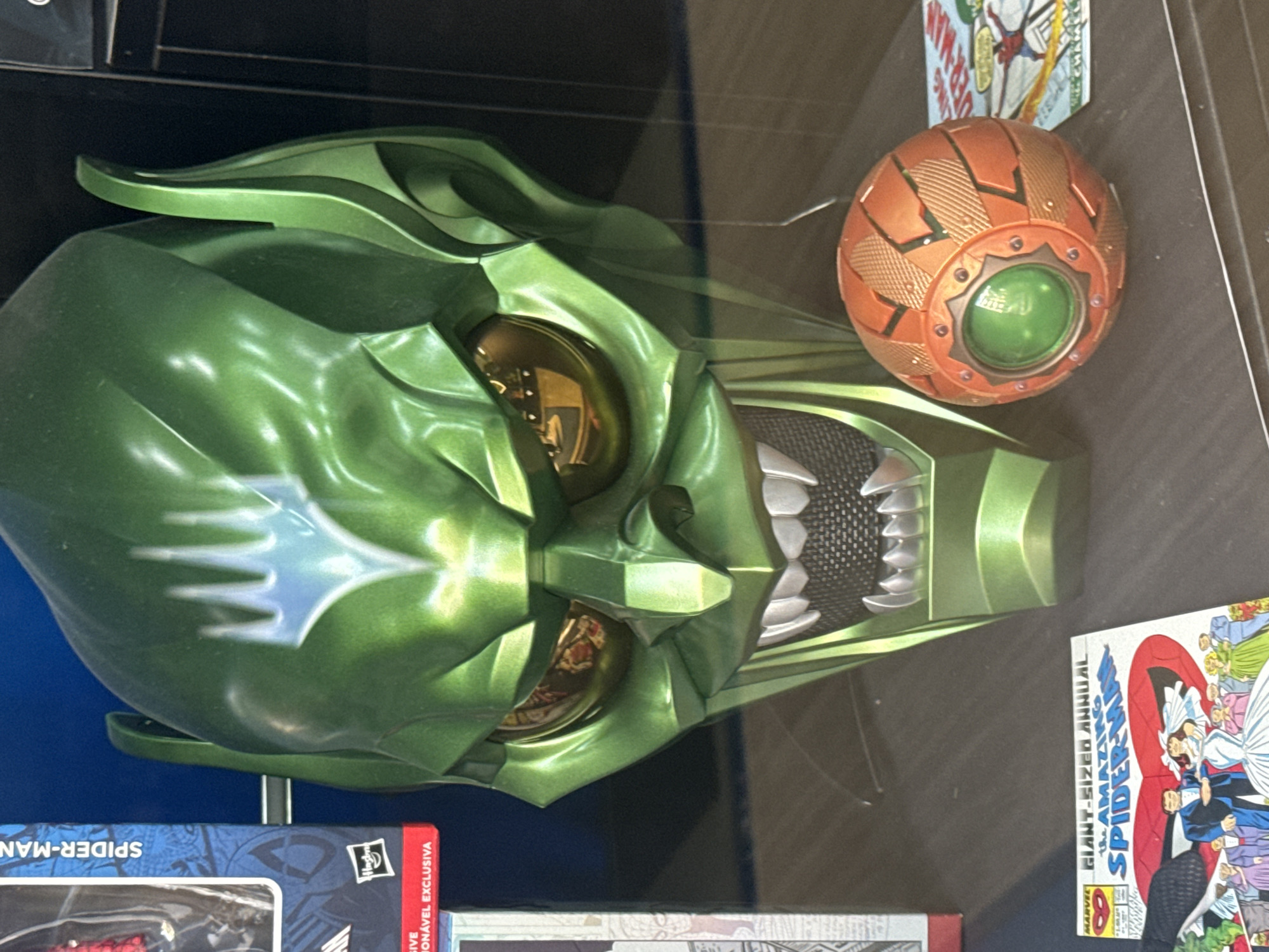 This image shows a Green Goblin mask and a pumpkin bomb, likely merchandise related to Spider-Man. The mask is predominantly green with silver accents and sharp, menacing teeth. The pumpkin bomb is orange with a green center. They appear to be displayed among other merchandise, possibly comics and action figures, some partially visible in the foreground and background. The setting seems to be indoors, possibly a retail environment or a collector's display. Location information (Barcelona, Spain) cannot be determined from the visual information available in the image.