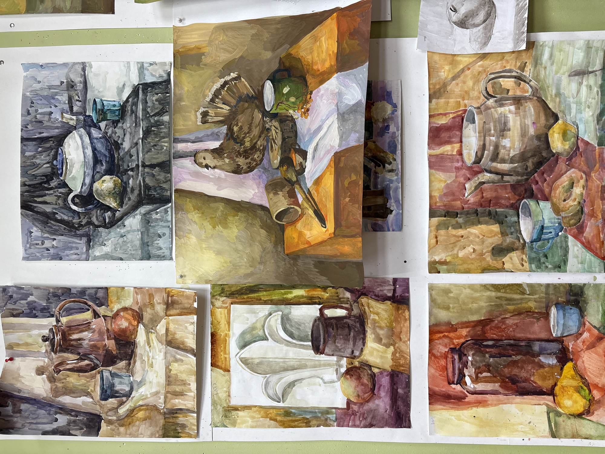 The image shows a collection of six watercolor paintings pinned to a wall. The paintings are still-life compositions of various objects. The objects depicted include teapots, cups, fruit (specifically pears and plums), jars, and a bird. One painting features the bird resting next to a green cup. The paintings are loosely rendered with varied color palettes. The wall is a light green. Some pinheads are visible, holding the paintings in place. The location is likely an art studio or classroom.