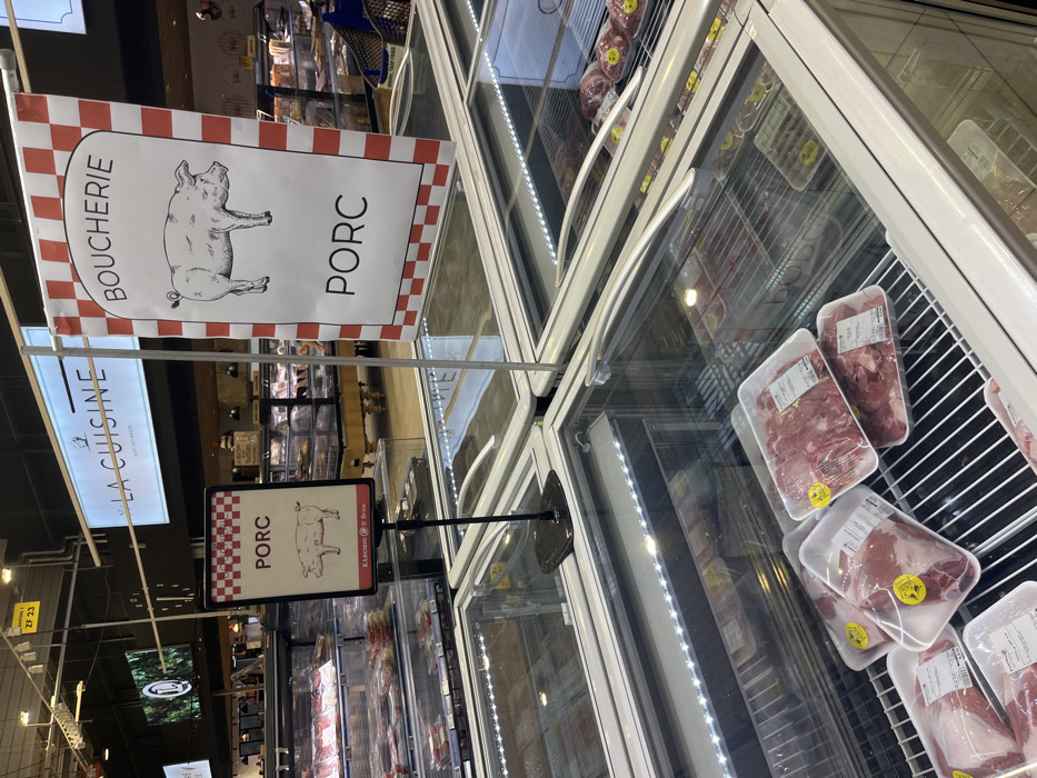 The image shows a refrigerated display case filled with packaged pork cuts in a supermarket in Saint-Brice-Courcelles, France (coordinates: 49.283, 3.999). Above the case, a sign reads "BOUCHERIE" with a cartoon pig and the word "PORC". Another sign above the case reads "LA CUISINE" and another, smaller sign reading "PORC" with a cartoon pig. All signs are in French. The case is made of glass and stainless steel. The lighting is fluorescent. The case is empty except for the packages of pork, which are displayed on metal shelves. The meat is raw and red. The packages are labeled with the word "PORC". The surrounding environment is a brightly lit supermarket.