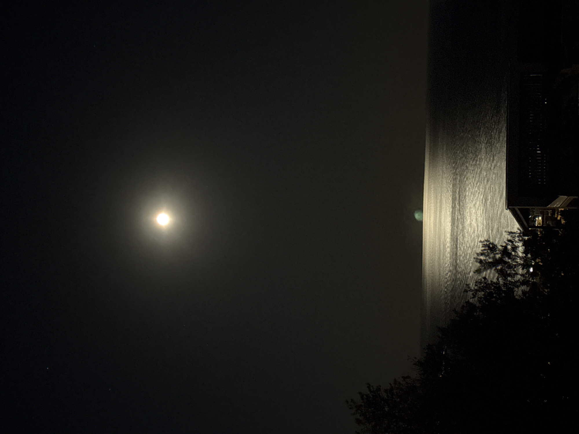 The image depicts a night scene with a bright light source, likely the moon, illuminating the dark sky. The light reflects off a body of water, creating a bright vertical line in the right portion of the image. A dark building or structure with visible architectural details is located on the right, partially obscured by trees or vegetation. The location is likely in Šušanj, Montenegro, as indicated in the prompt, and the scene appears to be near the coast, with the water reflecting the moonlight.