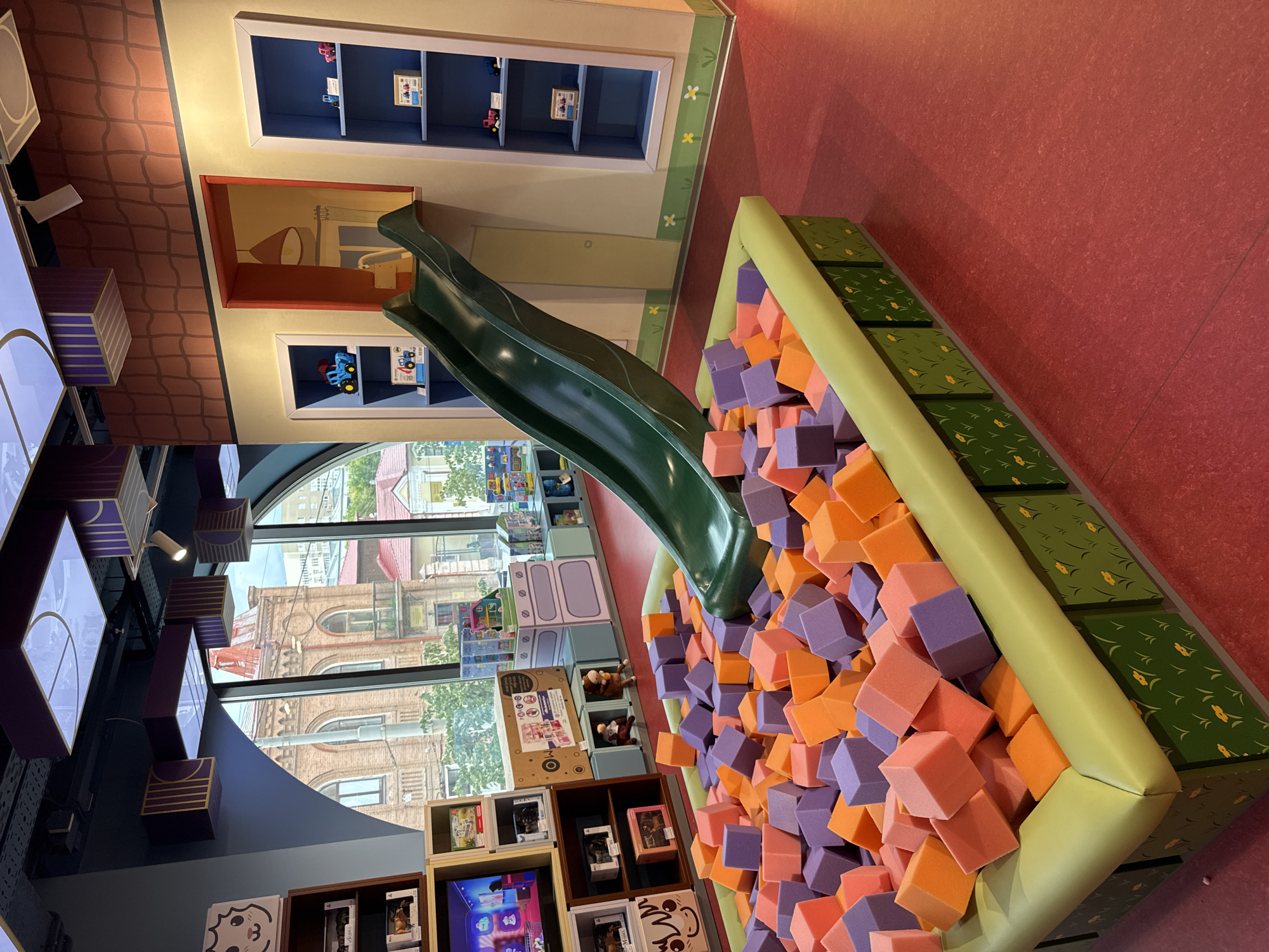 The image depicts an indoor play area, likely a children's entertainment center, with a focus on a soft play pit. The play pit is filled with numerous foam blocks in vibrant colors, including orange, purple, and pink. A green slide curves into the pit from an elevated position. The surrounding environment features brightly colored walls and shelving units displaying toys and children's books. Some shelves are designed to resemble dollhouses or miniature rooms. Through a large window, an exterior view shows buildings with classic architecture, suggesting an urban setting in Yekaterinburg, Russia. The light in the scene appears to be natural, possibly from an overcast day or late afternoon. The overall atmosphere is cheerful and playful, designed for children's recreation. There is no visible text in the image that can be read clearly.