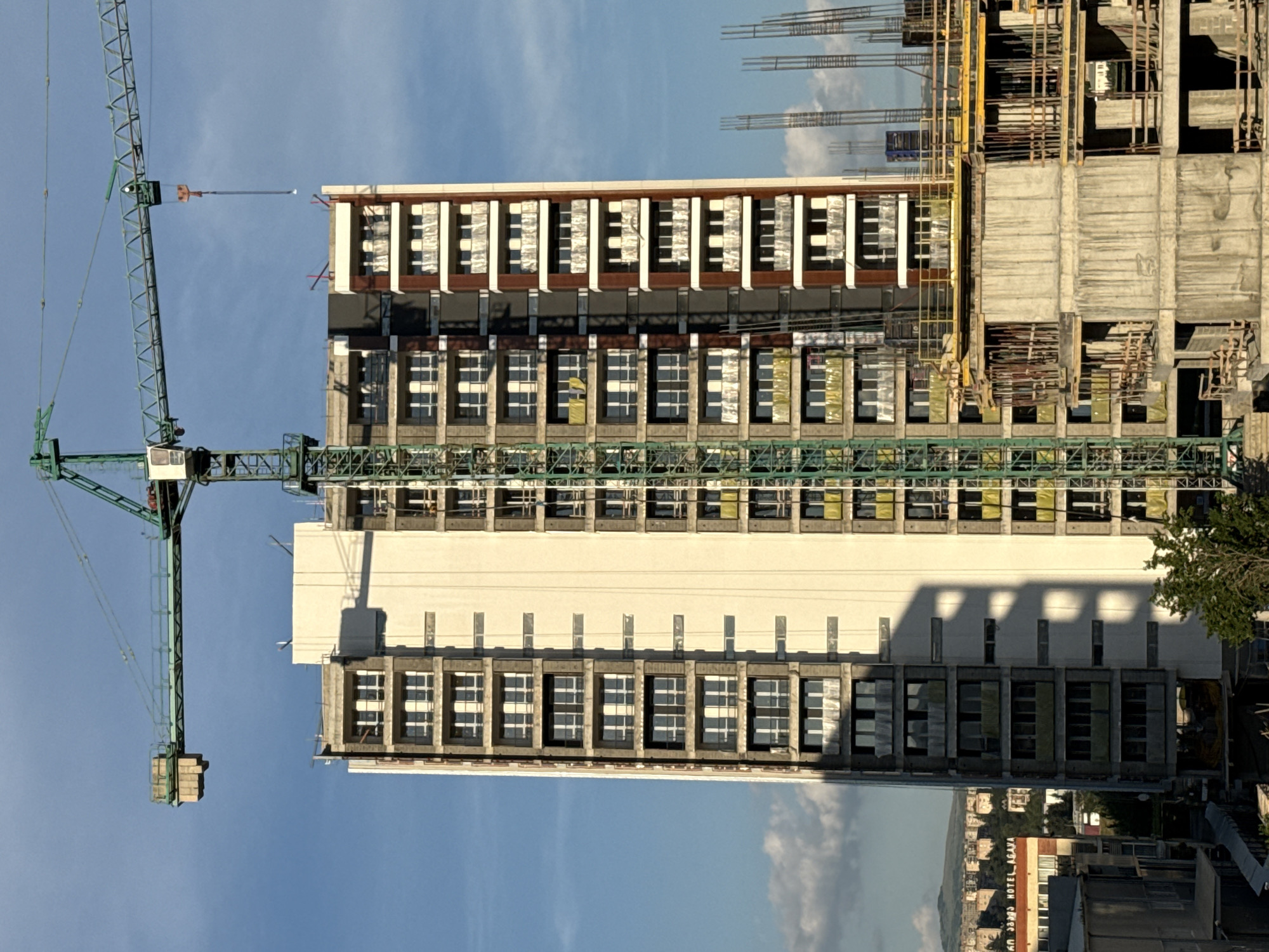 An exterior, eye-level, vertical medium shot captures a construction site in Tbilisi, Georgia, under a bright, hazy blue sky with sparse white clouds. Dominating the frame is a partially constructed tall building, its facade a mix of cream-colored panels and exposed concrete with numerous windows and balconies. A large green construction crane extends from the left side of the image, its boom casting a shadow on the building. In the foreground, another building under construction is visible, made of concrete with exposed rebar and scaffolding. In the background, a densely populated city area with smaller buildings and a distant, hazy mountain range can be seen. The lighting suggests it is late afternoon, with long shadows stretching across the scene.