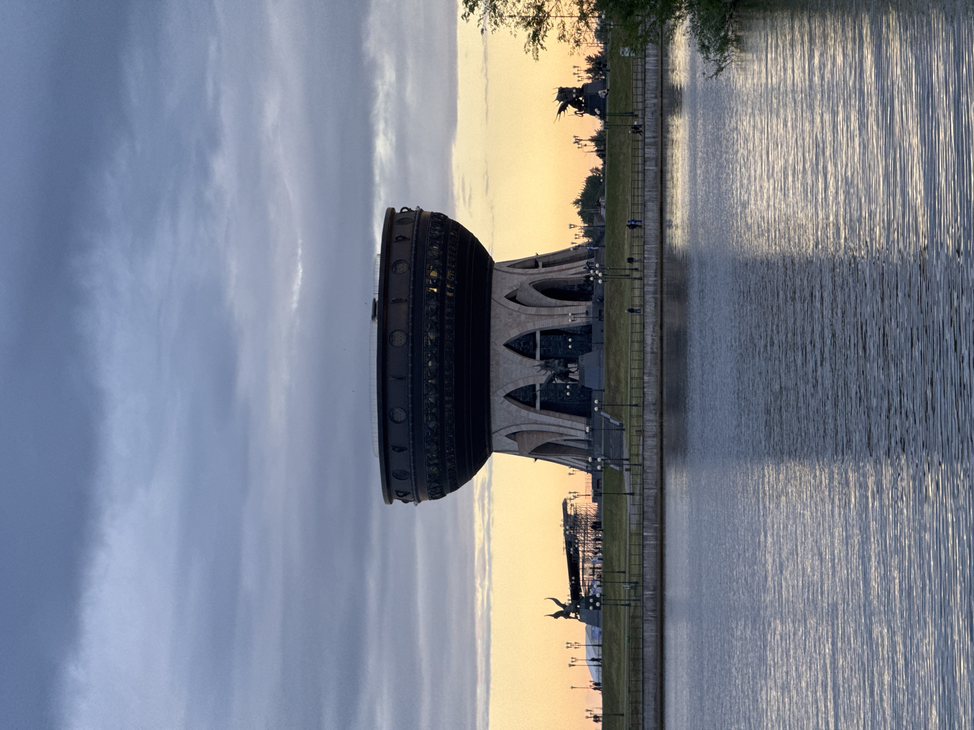 The image depicts the Kazan Family Center in Kazan, Russia, situated by the Kazanka River. The building, resembling a large cauldron or bowl, features intricate exterior detailing. Adjacent to the Family Center is a green lawn area interspersed with lampposts and sculptures, some of which are visible. The Kazanka River reflects the sky and surrounding structures. People are visible strolling along the riverbank. The sky transitions from a darker, cloudy sky to a lighter, yellow-tinged sky, possibly during sunrise or sunset.