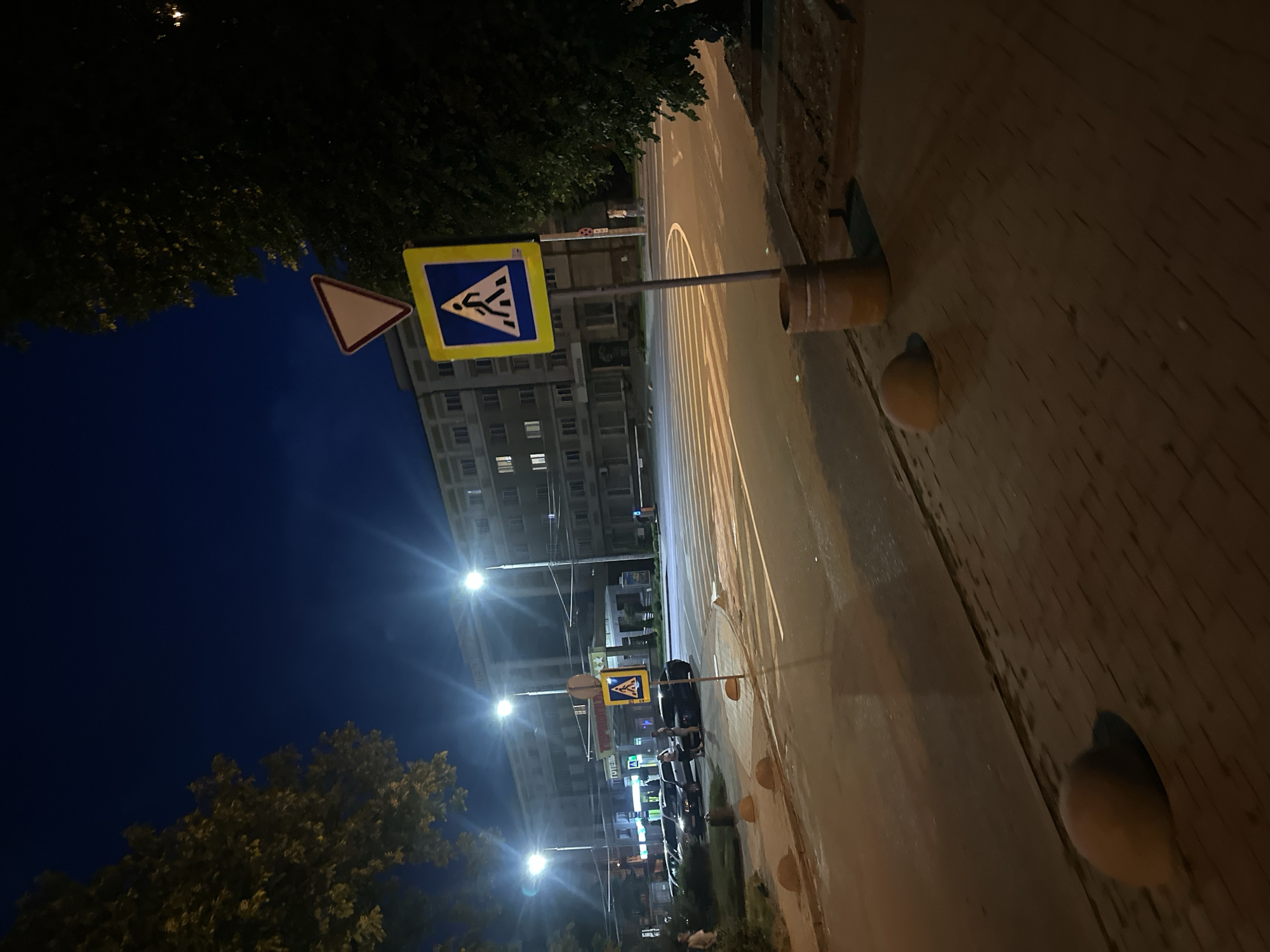 This nighttime image captures an urban street scene in Ternopil, Ukraine, centered around a pedestrian crossing. The sky is dark blue and clear. The foreground consists of a paved sidewalk with patterned pavers, featuring several hemispherical concrete bollards and a brown cylindrical trash receptacle. An asphalt road with clearly delineated white zebra stripes for a crosswalk stretches across the midground. Two identical blue square signs with yellow backgrounds depicting a white pedestrian symbol indicate the crossing; one is positioned on the right side of the road, topped by an inverted white triangle with a red border (yield sign), and another is visible further down on the left.
Several individuals are present: two people are standing near a dark sedan and a lighter car parked on the left side of the road, appearing to interact. Another person is seen walking in the distance on the far right. Multiple vehicles are parked along the left curb. The background is dominated by a multi-story building, identified as a hotel by the partially visible Cyrillic inscription "ГОТЕЛЬ" (Hotel) above its entrance. The scene is brightly illuminated by several streetlights on tall poles, casting strong light and producing lens flares. Trees with dark foliage frame both the left and right sides of the image.