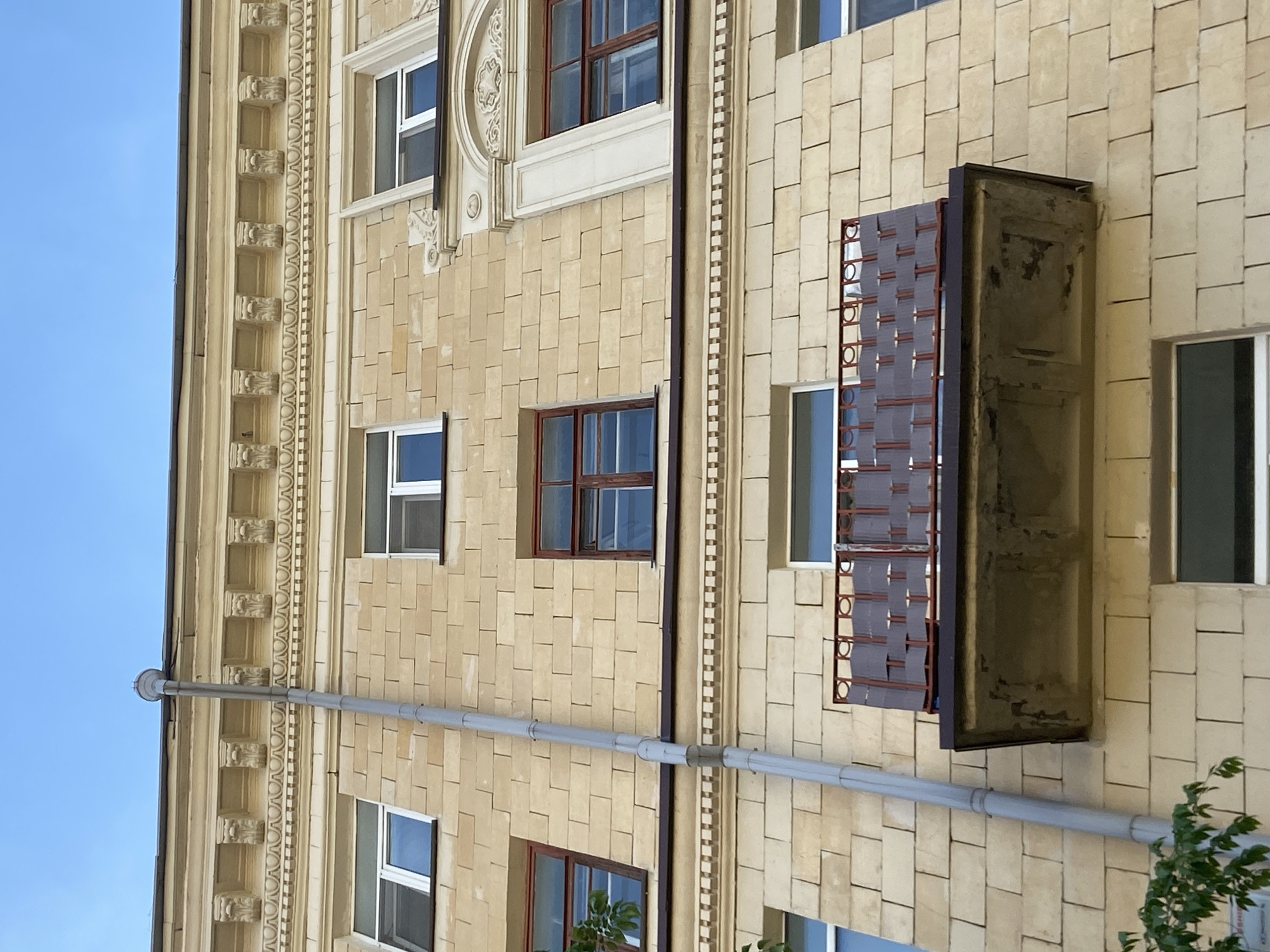 A slightly rotated, medium shot captures the facade of a building in Kazan, Russia, under a bright, clear sky. The building is constructed of light yellow, rectangular stones, arranged in a staggered pattern, giving a textured look. Several windows of varying sizes and trim styles are visible. Some have dark brown trim and others white, while some are adorned with decorative elements. A horizontal, gray pipe runs across the front of the building. A dark brown, box-like structure with a lattice covering extends outward from the facade, partially obstructing a window. The building is accented with ornate, light yellow architectural details running vertically along the side. A small green plant is visible at the bottom right of the shot.