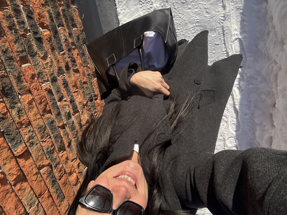 A selfie-style shot depicts a woman lying near a brick wall and snowy ground. She wears sunglasses and smiles, with her face close to the camera. A black coat and leather bag are beside her, and her dark hair is spread out around her head. The scene is brightly lit, suggesting an outdoor setting in daylight.