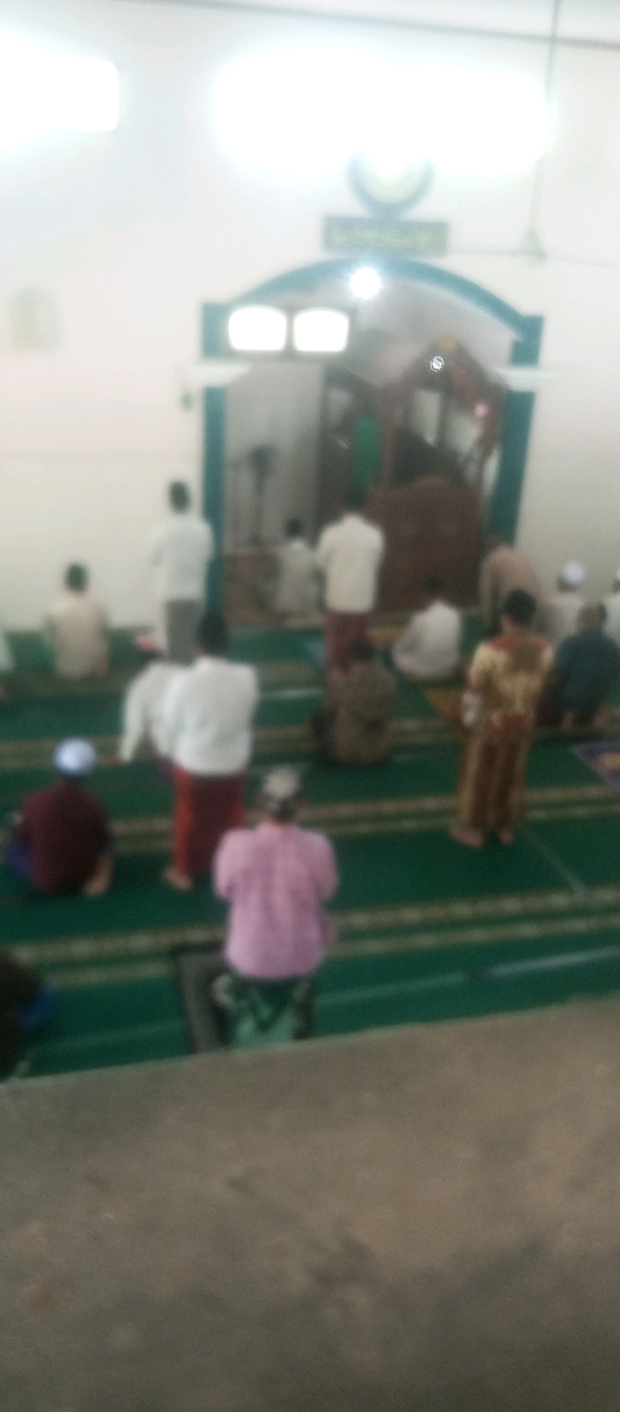 The image depicts a prayer gathering in a mosque, likely in Kediri City, Indonesia, given the context provided. The scene is characterized by a group of people, mostly men, engaged in Islamic prayer, indicated by their kneeling and standing postures on a green carpet with striped markings. The lighting is bright, creating a somewhat overexposed effect, and the image is slightly blurred, obscuring fine details of the individuals and their clothing.
In the foreground, a person is seen kneeling, facing away from the camera. Further back, several individuals are positioned in prayer rows. Some are standing, with their hands raised, a common stance during prayer, while others are in a kneeling or prostrating position. The environment appears to be an indoor prayer hall with a white wall and a ceiling. A raised wooden structure, possibly a pulpit or a platform for religious leaders, is visible in the background, with an arched entrance. There is an ornamental element above the arch. The overall atmosphere suggests a religious service or gathering within a mosque. The presence of a fan in the background hints at the functional aspects of the space. The image's blurriness and bright lighting make it difficult to ascertain specific demographic details or individual identities.