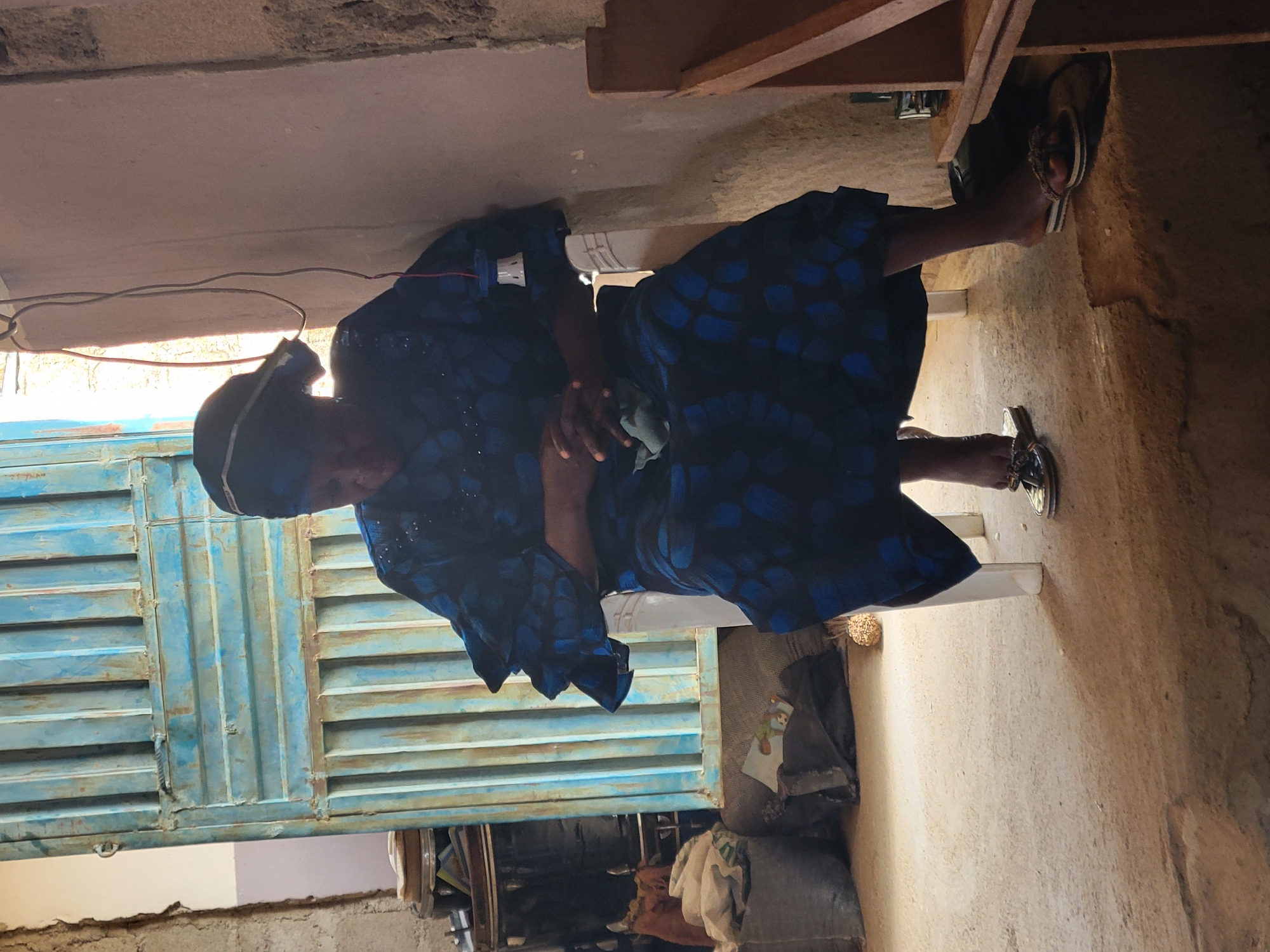 In Madalla, Nigeria, a woman wearing a dark blue dress with a blue floral pattern sits on a white plastic chair in an indoor setting. Her hands are clasped in her lap, and she is wearing a dark headscarf. The environment appears to be a room with a light-colored, textured wall on the right and a closed, blue, corrugated metal door on the left. In the background, stacks of items are visible, including large grey bags and what appear to be drums or barrels. A light source shines in from the right, casting shadows on the floor. The floor is made of rough, uneven concrete. The woman is wearing open-toed sandals.