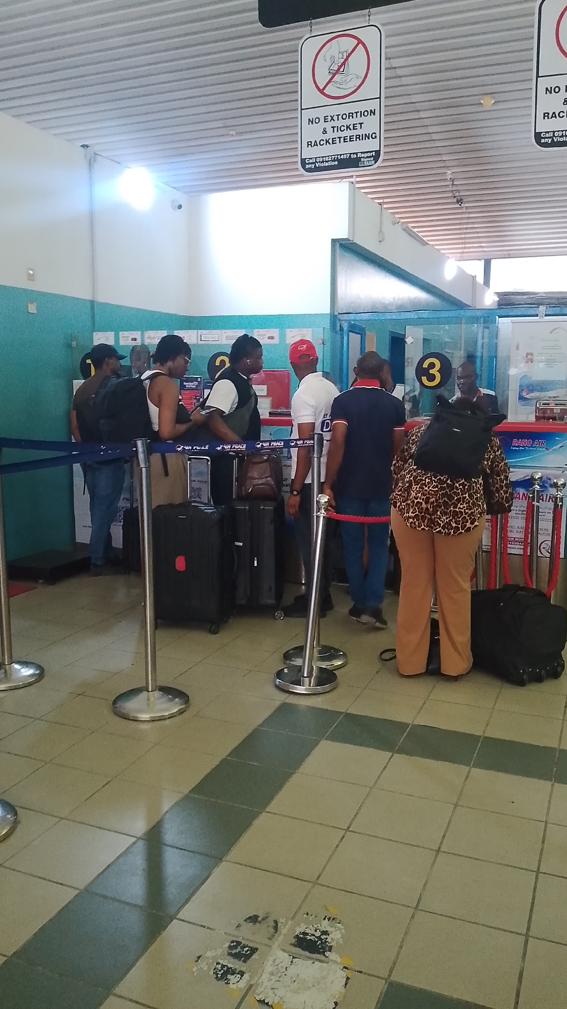The photo shows people lining up at a ticket counter in what appears to be an airport or similar transportation hub. The location is suggested to be Orerokpe, Nigeria.
Several individuals stand in a queue delineated by stanchions with blue ropes. Luggage is scattered amongst the people. A sign overhead reads "NO EXTORTION & TICKET RACKETEERING" and provides a number to report violations. Behind the counter, a number 3 is visible above a person attending to the line. The airline logo and the names of several towns is visible below the counter window. The floor consists of square tiles, some of which show wear and stains.